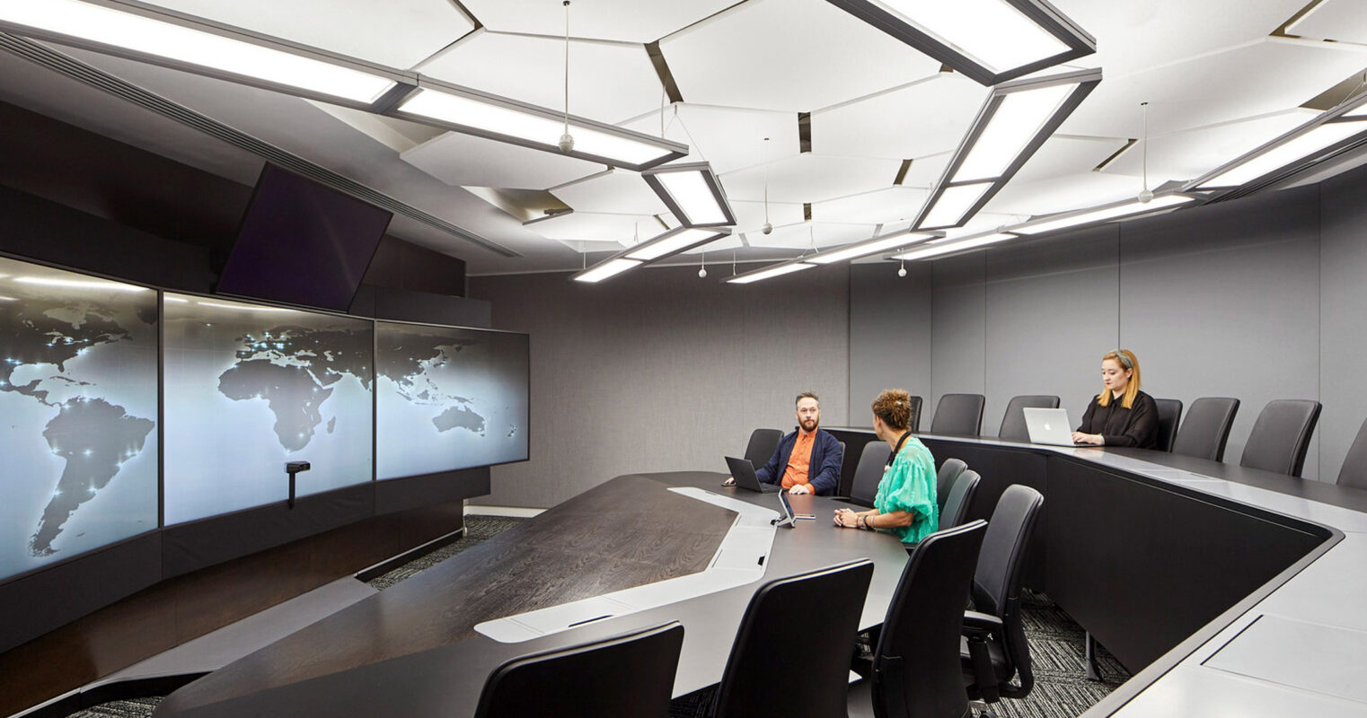Modern conference room featuring an angular black table, a world map on digital screens along one wall, and geometric ceiling lighting, with three individuals engaged in a discussion.
