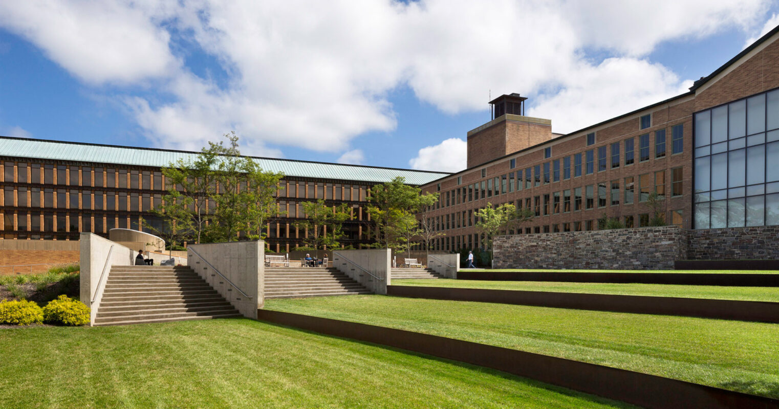 Broad lawn flanked by tiered landscaped areas in front of an expansive modern brick building with large glass windows, featuring outdoor seating and pedestrians.