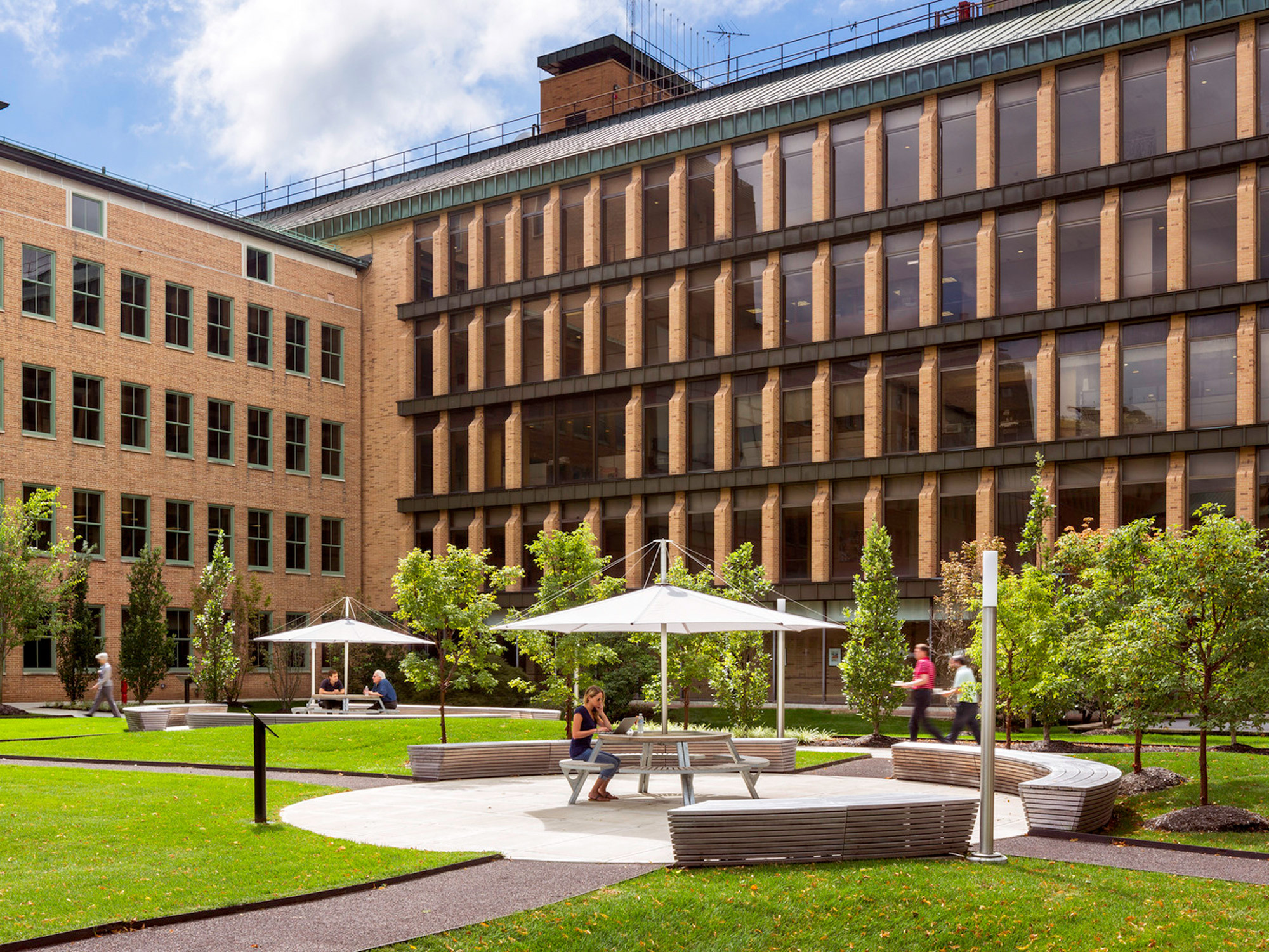 Modern university courtyard showcasing a blend of natural and architectural design, with students occupying benches and tables beneath white parasols, framed by a lush green lawn and a multi-story academic building with brick and glass facade.