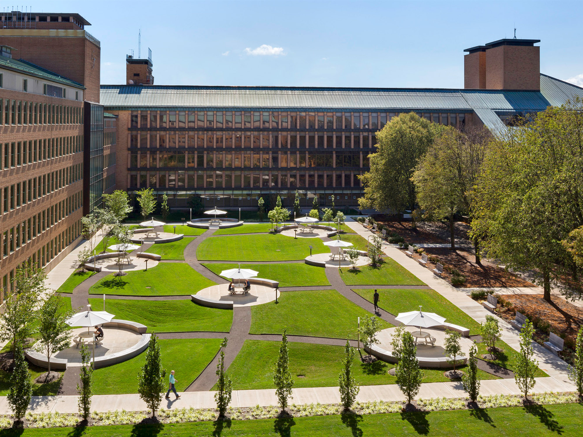 Lush green lawn with symmetrical garden beds and circular seating areas punctuates the courtyard, surrounded by a cohesive blend of modern and traditional brick architecture under a clear blue sky.