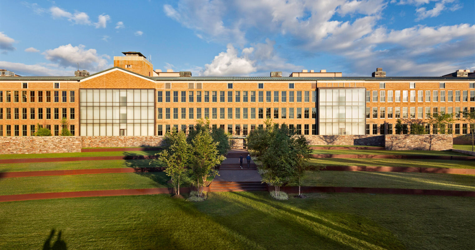 Large contemporary educational building with symmetrical design, expansive glass windows, and a flat roof. A central clock tower stands above an articulated brick facade, bordered by a manicured lawn and pathways in a campus setting at golden hour.