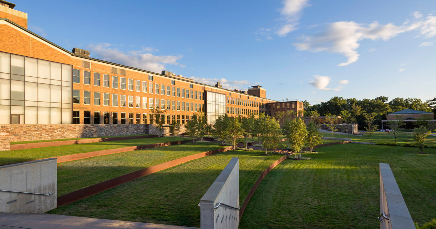 Modern educational facility with a blend of traditional brickwork and contemporary glass facade, landscaped with green lawns, geometric walkways, and young trees, under a clear blue sky.