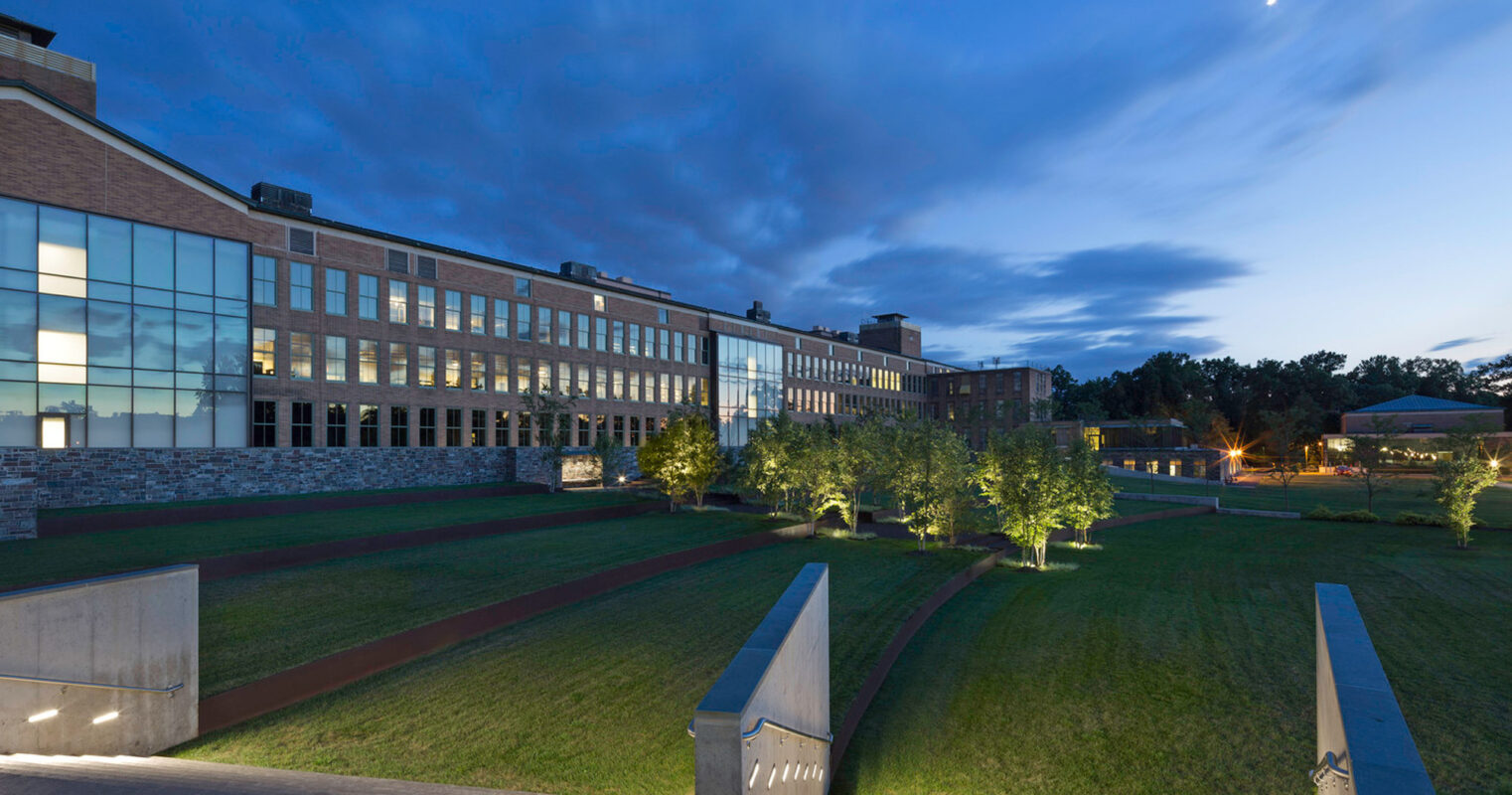 Twilight view of a modern educational building with expansive glass facade, illuminated interior rooms, stone accents, and a manicured lawn with young trees. Sleek benches and concrete pathways enhance the functional aesthetic of the outdoor space.