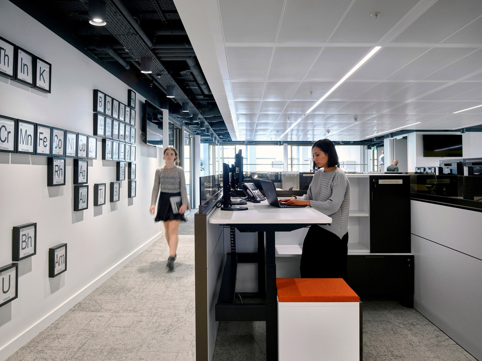 A modern office space with an open floor plan featuring a standing work station, periodical table-themed wall art, and sleek, monochromatic color scheme. Employees engage in a productive, contemporary environment.