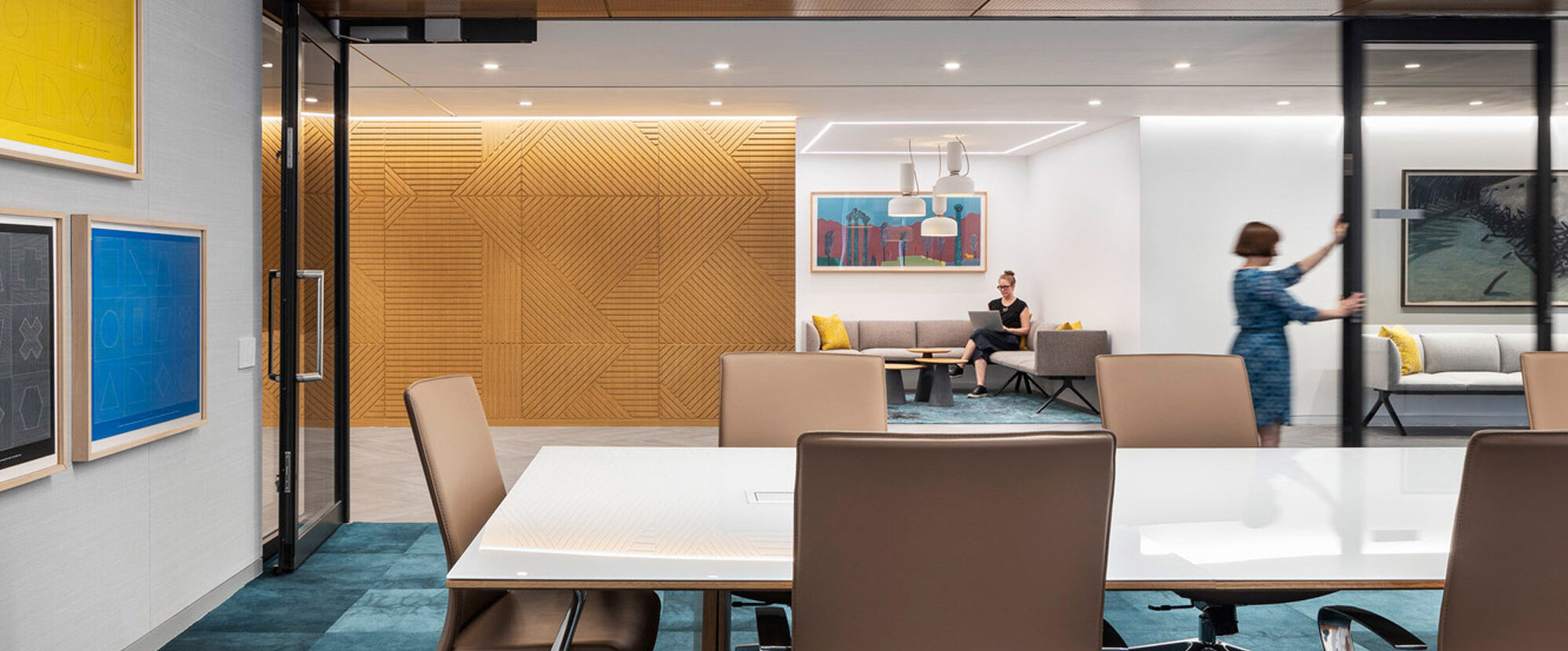 Modern office conference room featuring a sleek rectangular table, leather rolling chairs, and wood-paneled walls with integrated lighting. Vibrant artwork and deep blue carpet add a pop of color, while glass walls provide transparency and connectivity to adjacent spaces.