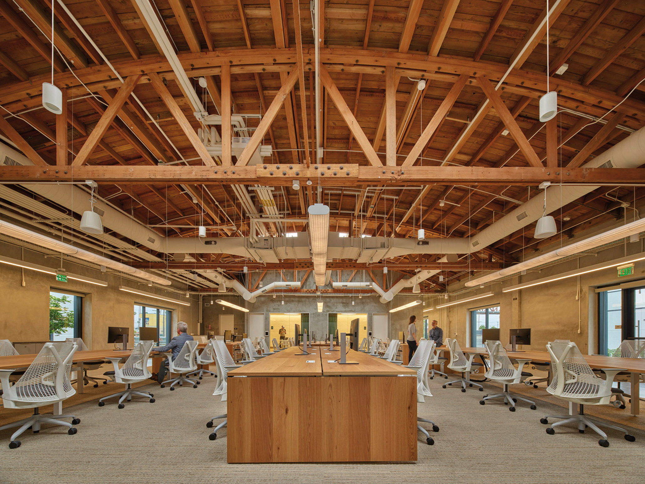Modern office space featuring exposed wooden truss ceilings and pendant lighting. Central communal workstation surrounded by individual desks, ergonomic chairs, and ample natural light.