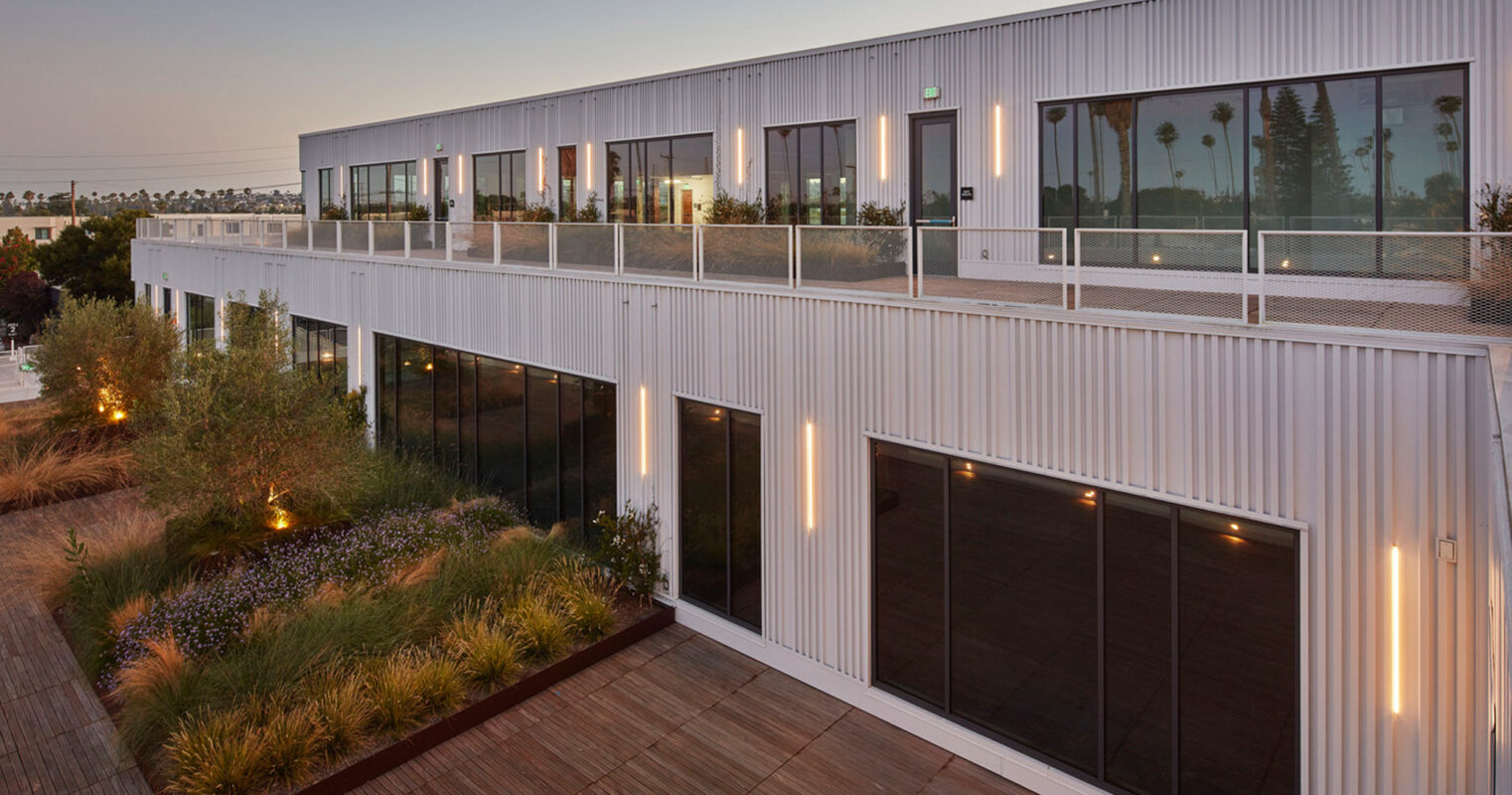 Modern industrial building exterior at dusk with ribbed metal cladding, floor-to-ceiling windows, and integrated planters on a rooftop terrace with ambient lighting.