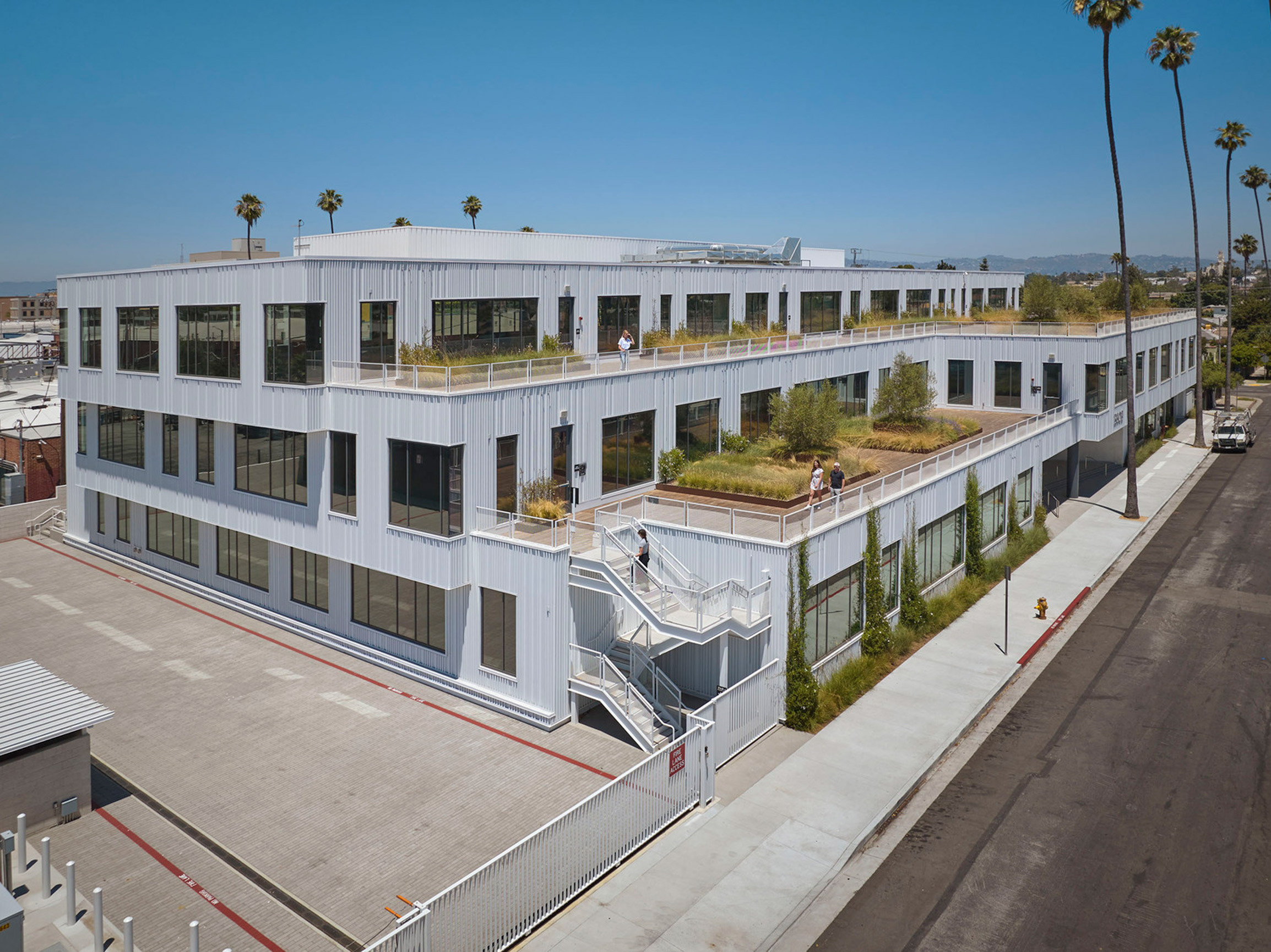 Modern two-story office building with asymmetrical design featuring a white facade, expansive windows for natural lighting, and eco-friendly green roof terraces amidst an urban landscape with palm trees.