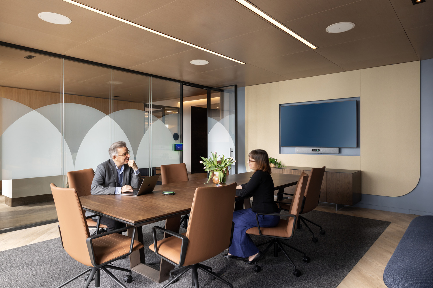 Modern office meeting room with a wooden table, leather chairs, frosted glass partitions, and wooden floor. A flat-screen monitor is mounted on the wall, and two professionals are in discussion, embodying a collaborative workspace aesthetic.