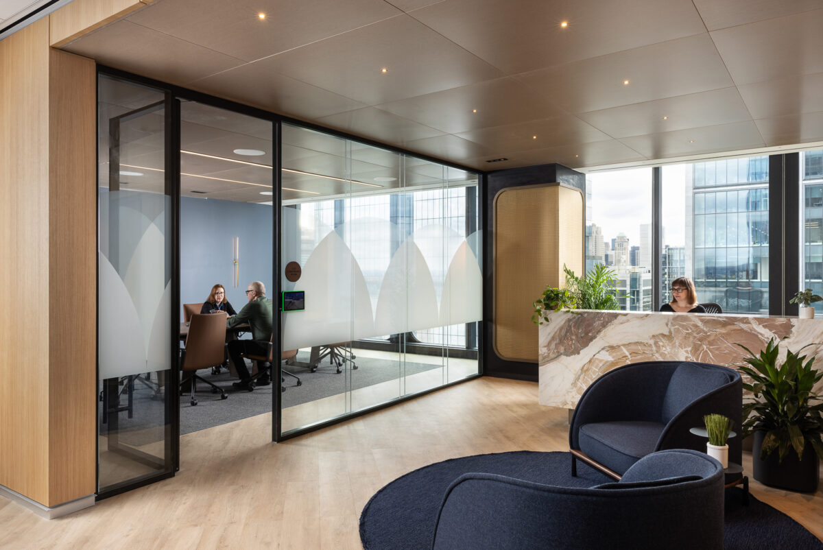 Modern office space featuring sleek glass partitions with frosted geometric patterns, navy blue armchairs, light wood flooring, and potted greenery enhancing a warm yet corporate atmosphere.