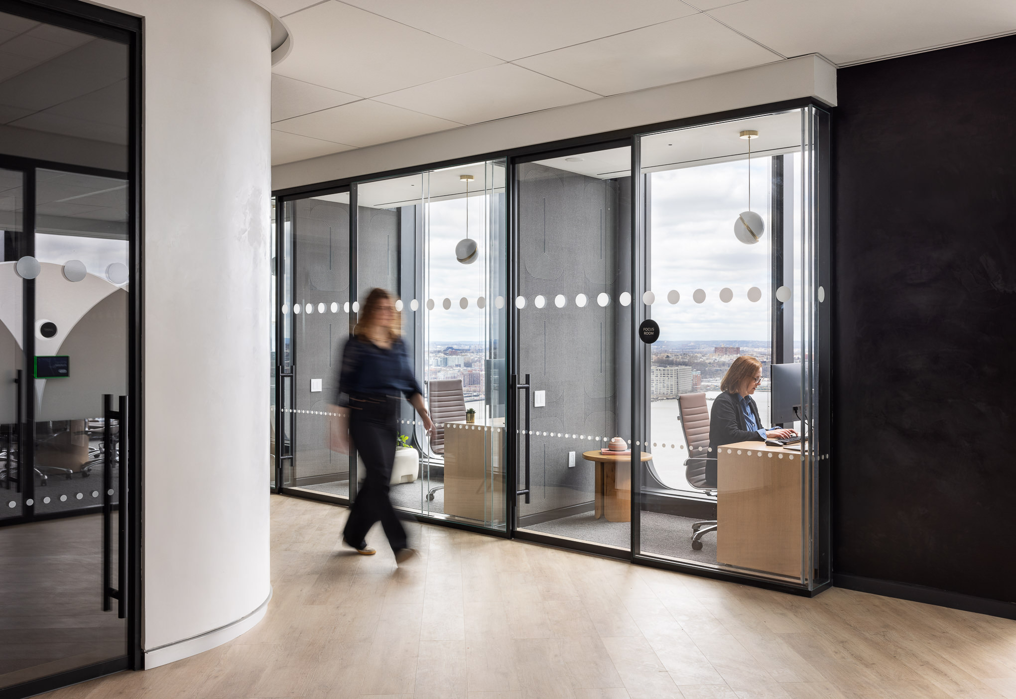 Modern office space with floor-to-ceiling glass partitions maximizing natural light. The interior showcases minimalistic furniture, neutral tones, and accent lighting, blending functionality with a contemporary aesthetic. Reflections hint at a high-rise setting, indicating an urban context for this professional environment.
