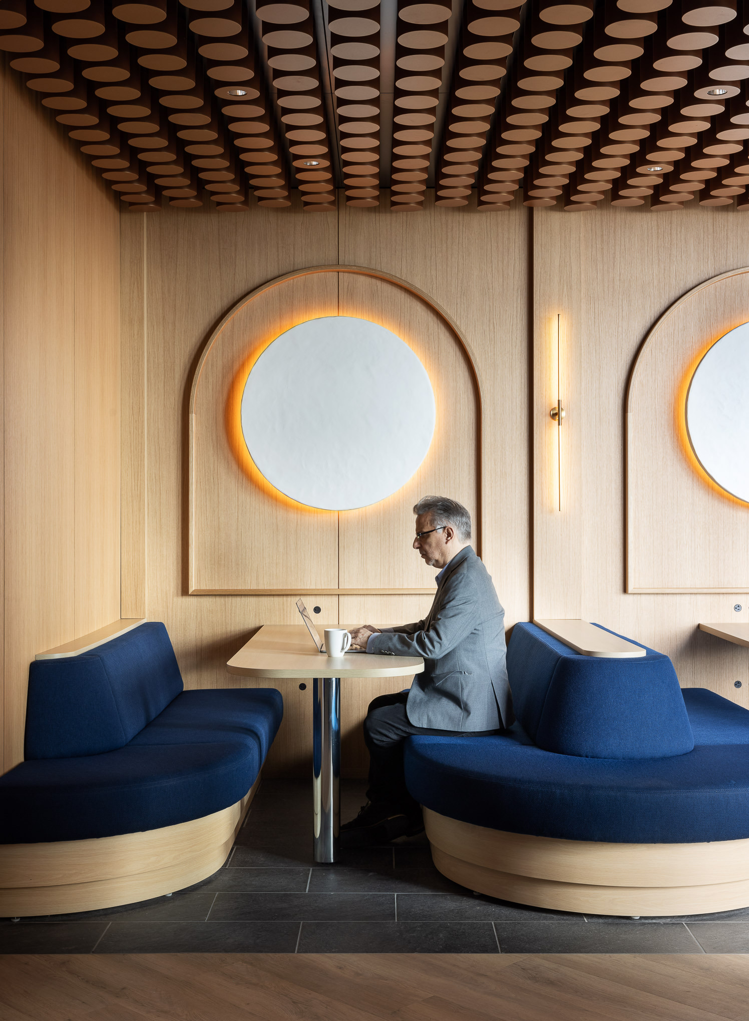 Elegant cafe interior featuring curved dark blue banquettes and warm wooden wall panels with circular cutouts and backlit accents. A gentleman in a grey suit works at a round table, highlighting the space's modern yet inviting ambiance.