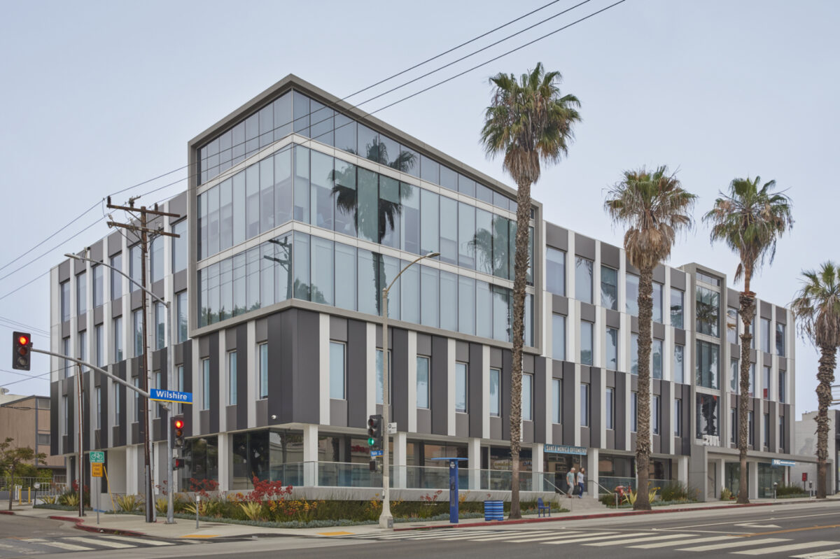Modern mixed-use building featuring a striking geometric facade with a combination of glass and textured materials. Palm trees line the entrance, enhancing the urban landscape. Large windows promote natural light, while the contemporary design integrates seamlessly with its surroundings, reflecting a progressive approach to urban architecture.