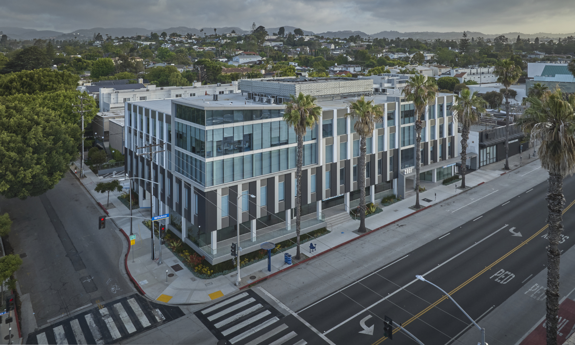 Modern commercial building featuring a sleek, geometric façade with alternating dark and light tones. Large glass windows reflect the surroundings, enhancing natural light. Palm trees line the street, contributing to a vibrant, urban atmosphere, while the structure’s design emphasizes functionality and contemporary architectural aesthetics.