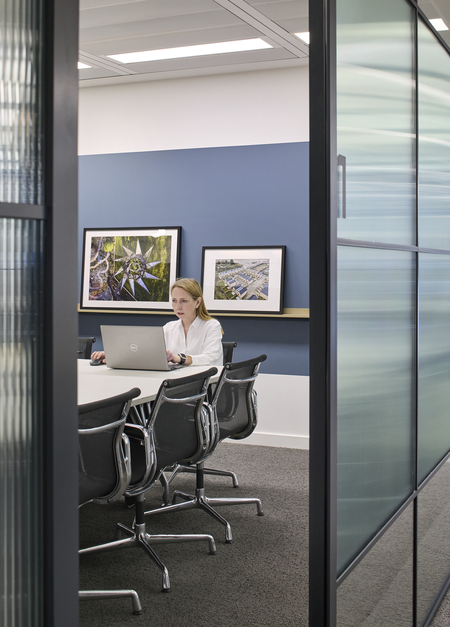 A modern conference room features a sleek, glass wall and minimalist design. A woman in a white blouse works focused at a laptop in front of a large table surrounded by ergonomic chairs. Framed artworks adorn the blue accent wall, adding a touch of creativity to the professional space.