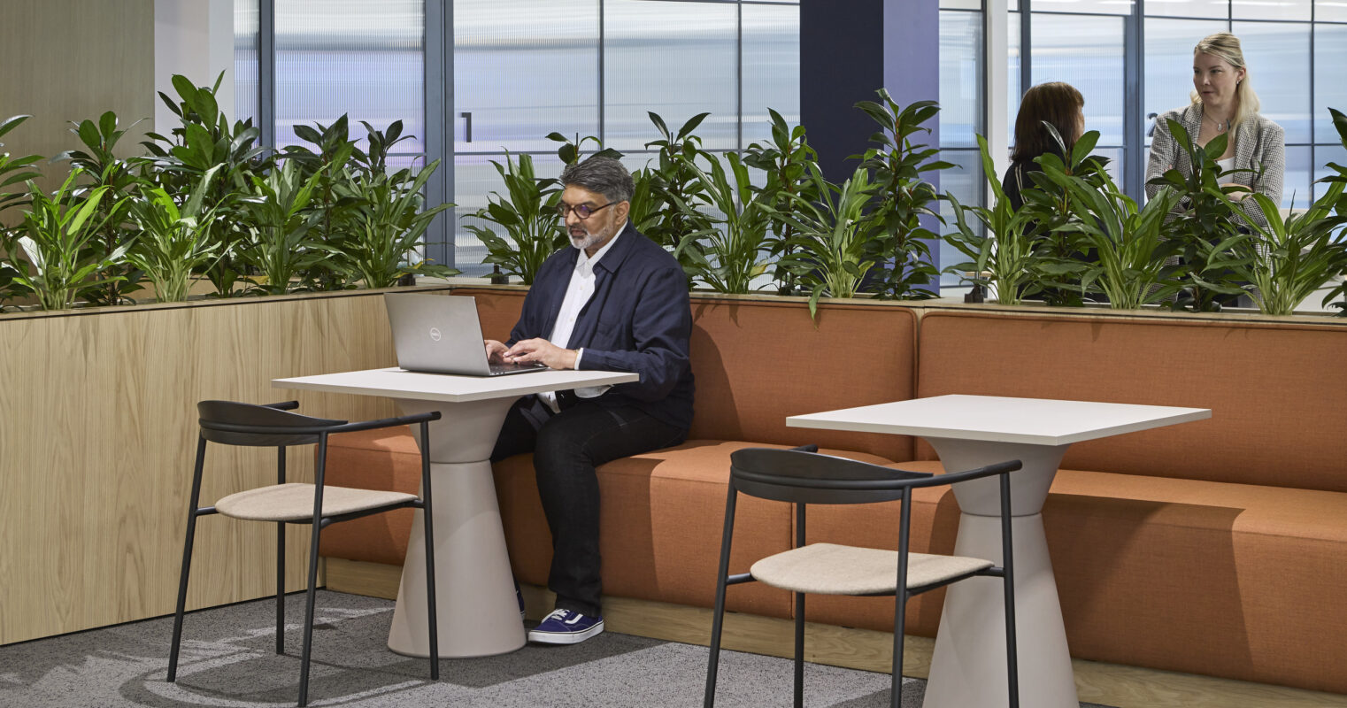 A modern workspace features a man using a laptop while seated at a sleek table on a textured carpet. Surrounding greenery enhances the inviting atmosphere, complemented by warm orange seating. The open layout supports collaboration, with additional team members visible in the background, showcasing a blend of nature and contemporary design.