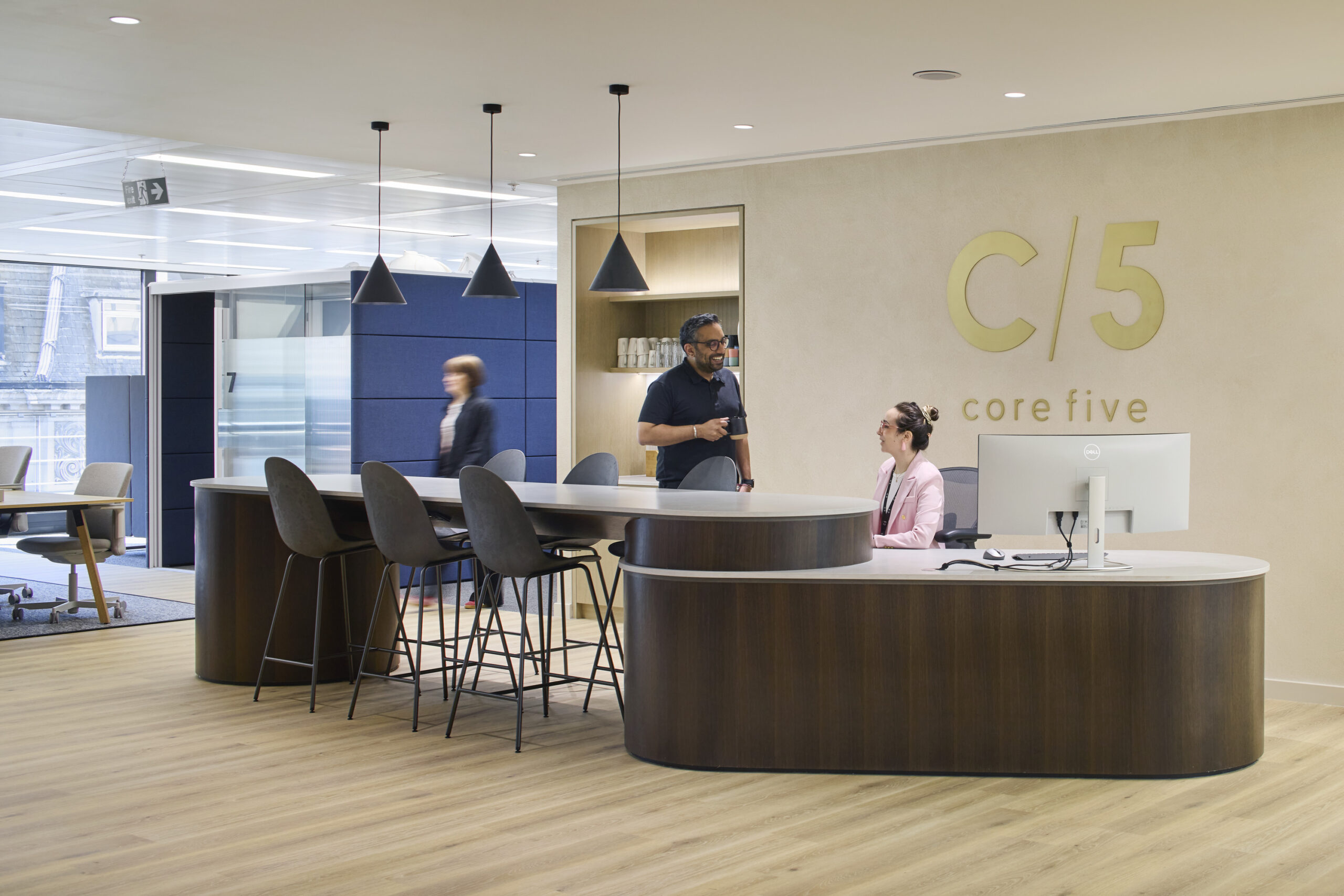 A modern reception area featuring a sleek curved reception desk crafted from warm wood. Stylish pendant lights hang above, illuminating the space. A comfortable seating arrangement, including high stools and chairs, invites collaboration. The neutral color palette and large windows create an open, inviting atmosphere, promoting productivity and comfort.