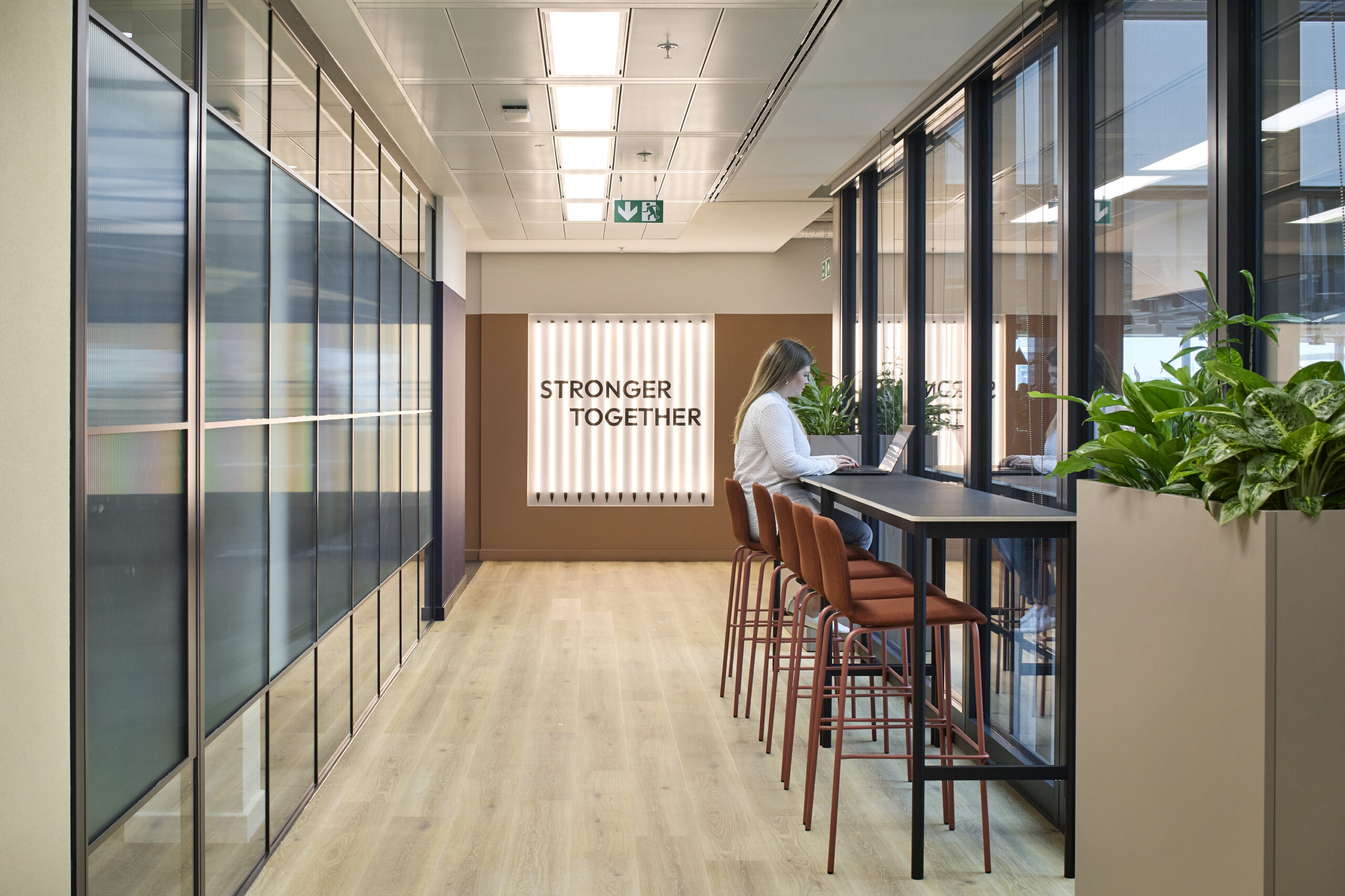 Bright and open office space featuring a modern design with large glass partitions. A stylish communal table lined with brown, upholstered bar stools invites collaboration. Natural light filters through the glass, highlighting the "STRONGER TOGETHER" sign on the wall and lush greenery in planters, promoting a welcoming atmosphere.