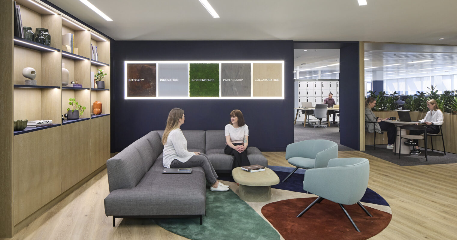In a modern office lounge, two professionals engage in conversation on a contemporary gray sofa. The space features a stylish circular rug with earth-tone accents and a backdrop of wooden shelves displaying decorative items. Colorful framed words emphasize core values, enhancing the inviting ambiance for teamwork.
