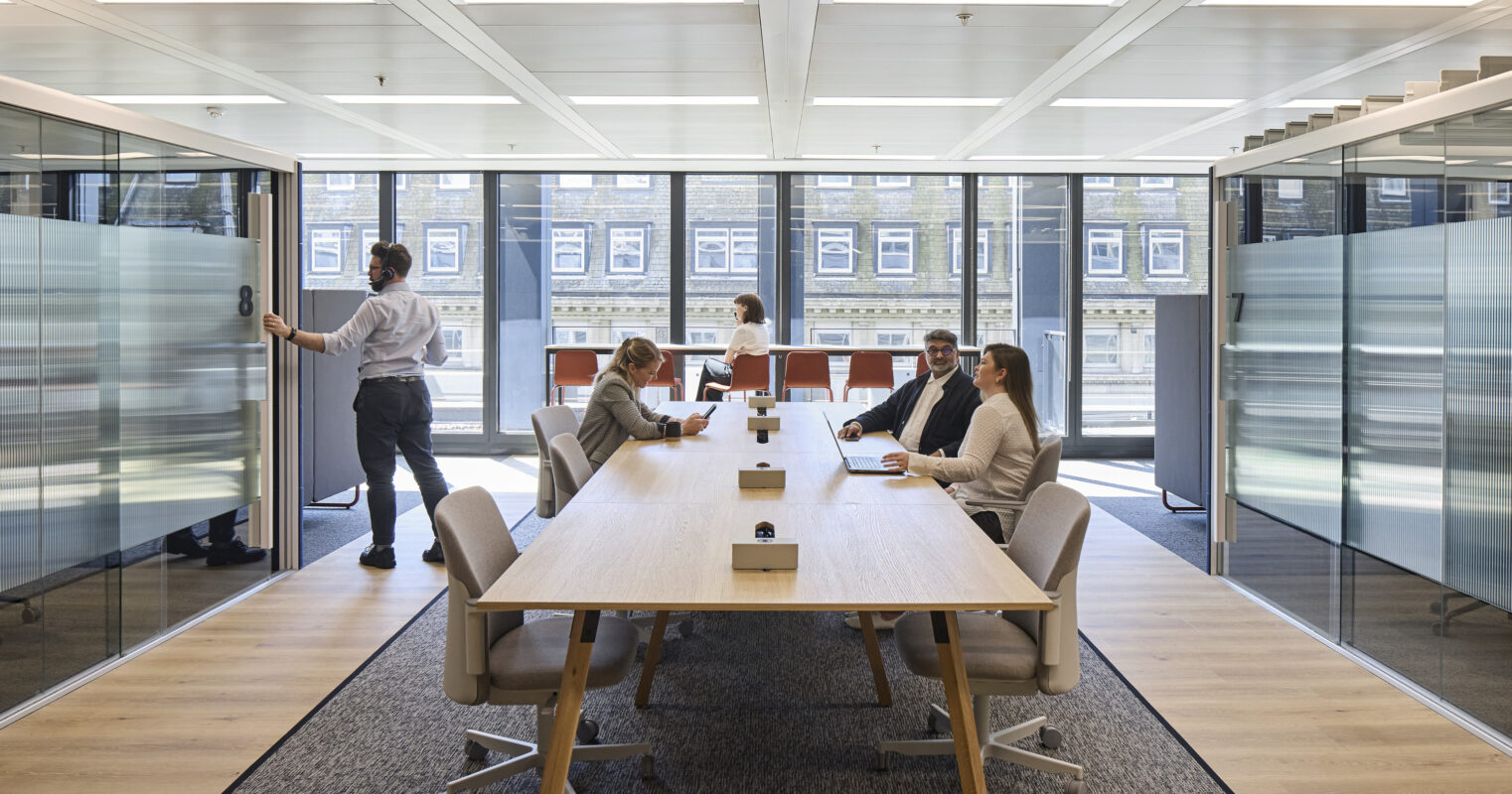 A modern conference room featuring a long wooden table surrounded by ergonomic chairs, illuminated by natural light from large windows. Glass partition walls create an open feel, while stylish patterned carpet complements the minimalist decor. Professionals engage in discussions, emphasizing collaboration and contemporary design.