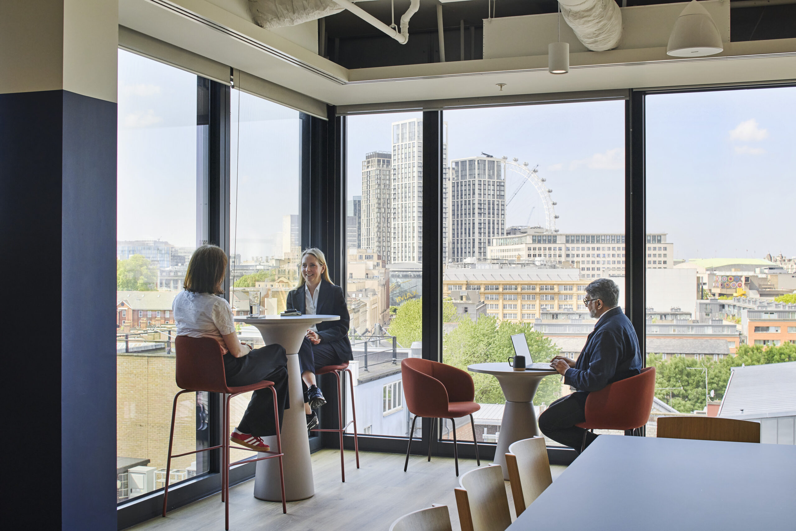 Contemporary open workspace featuring a panoramic view of London, showcasing a mix of modern furnishings. High tables with stools create a collaborative atmosphere, while large windows flood the space with natural light. The design emphasizes functionality and comfort, blending urban aesthetics with a productive work environment.