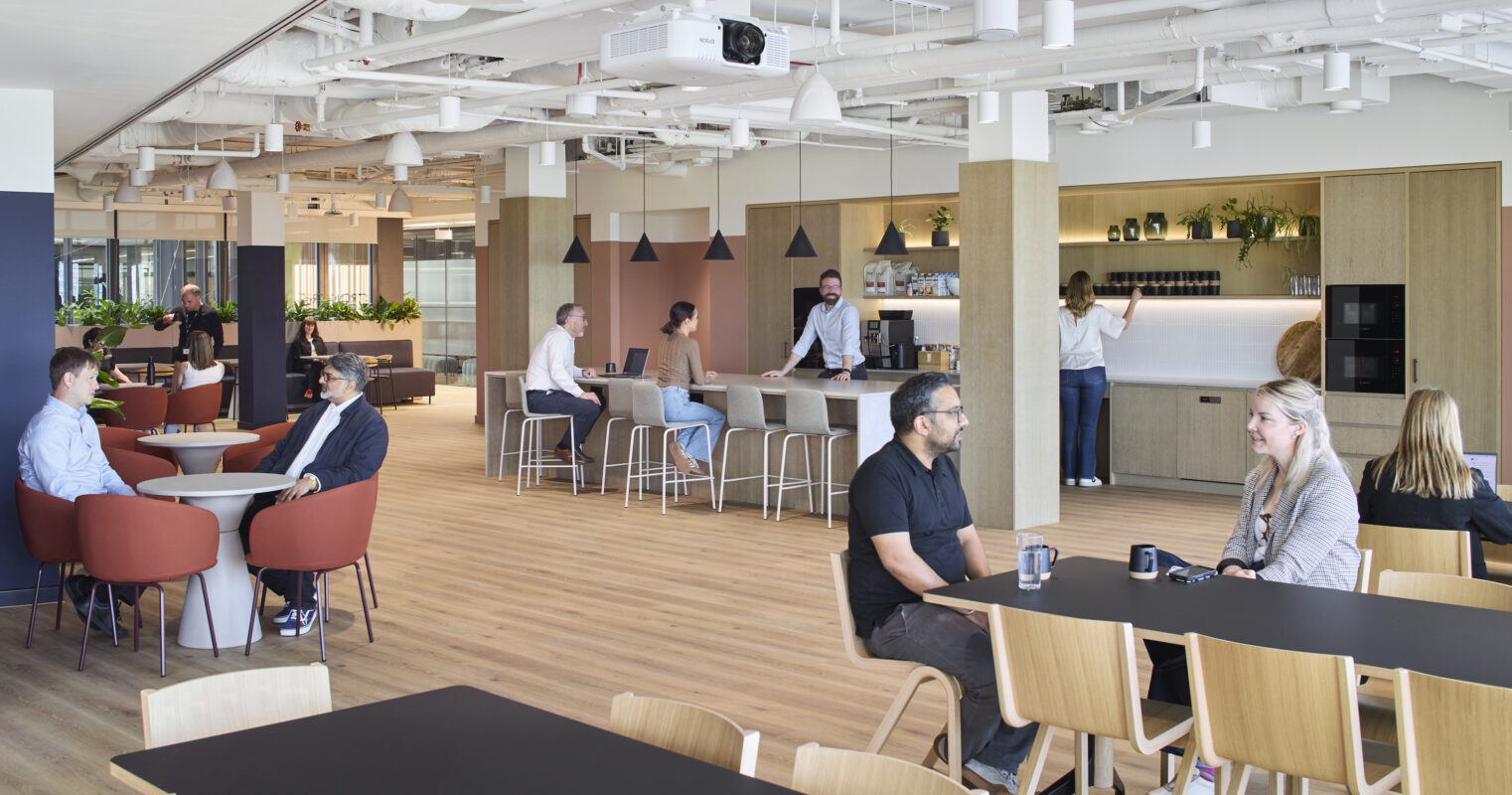 A contemporary office space featuring an open layout with various seating arrangements. Groups of professionals engage in conversation around circular and rectangular tables. A modern kitchenette with sleek cabinetry and plants creates a welcoming atmosphere, complemented by warm wooden flooring and bright lighting, enhancing collaboration and productivity.