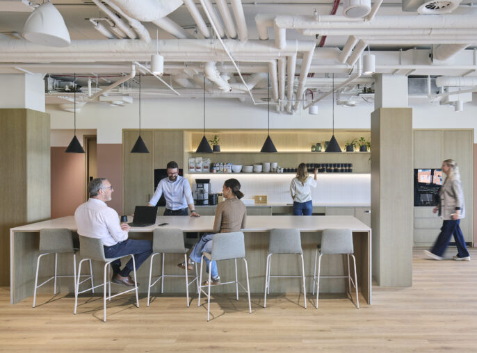 A modern office kitchen area features a minimalist design with natural wood cabinetry and sleek countertops. A communal table with high stools invites collaboration, while pendant lights add a warm ambiance. Light-filled space enhances productivity, showcasing a harmonious blend of functionality and aesthetics in contemporary workplace design.