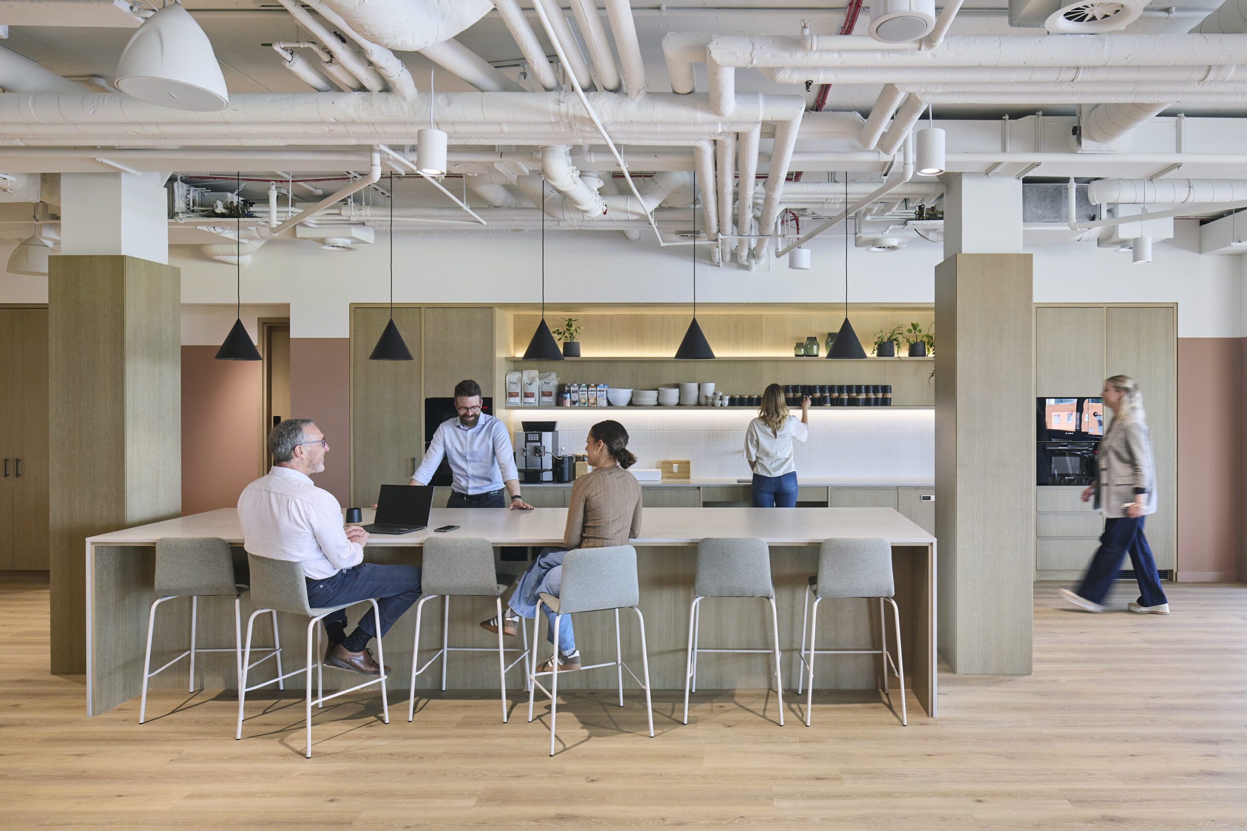 A modern office kitchen area features a minimalist design with natural wood cabinetry and sleek countertops. A communal table with high stools invites collaboration, while pendant lights add a warm ambiance. Light-filled space enhances productivity, showcasing a harmonious blend of functionality and aesthetics in contemporary workplace design.