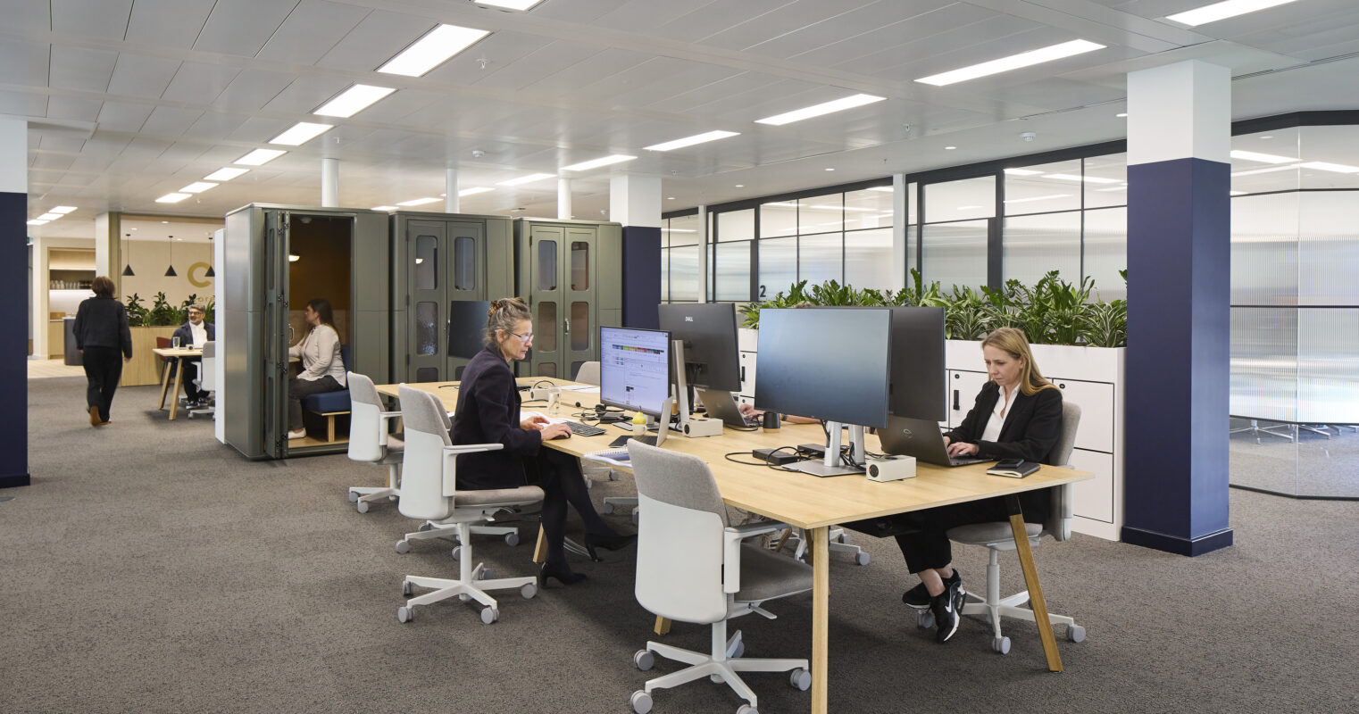 A modern office interior featuring an open-plan layout with collaborative workspaces. Two employees are engaged at sleek desks equipped with dual monitors, while others utilize private booths in the background. Abundant natural light floods through large windows, complemented by plants that enhance the workspace's biophilic design.