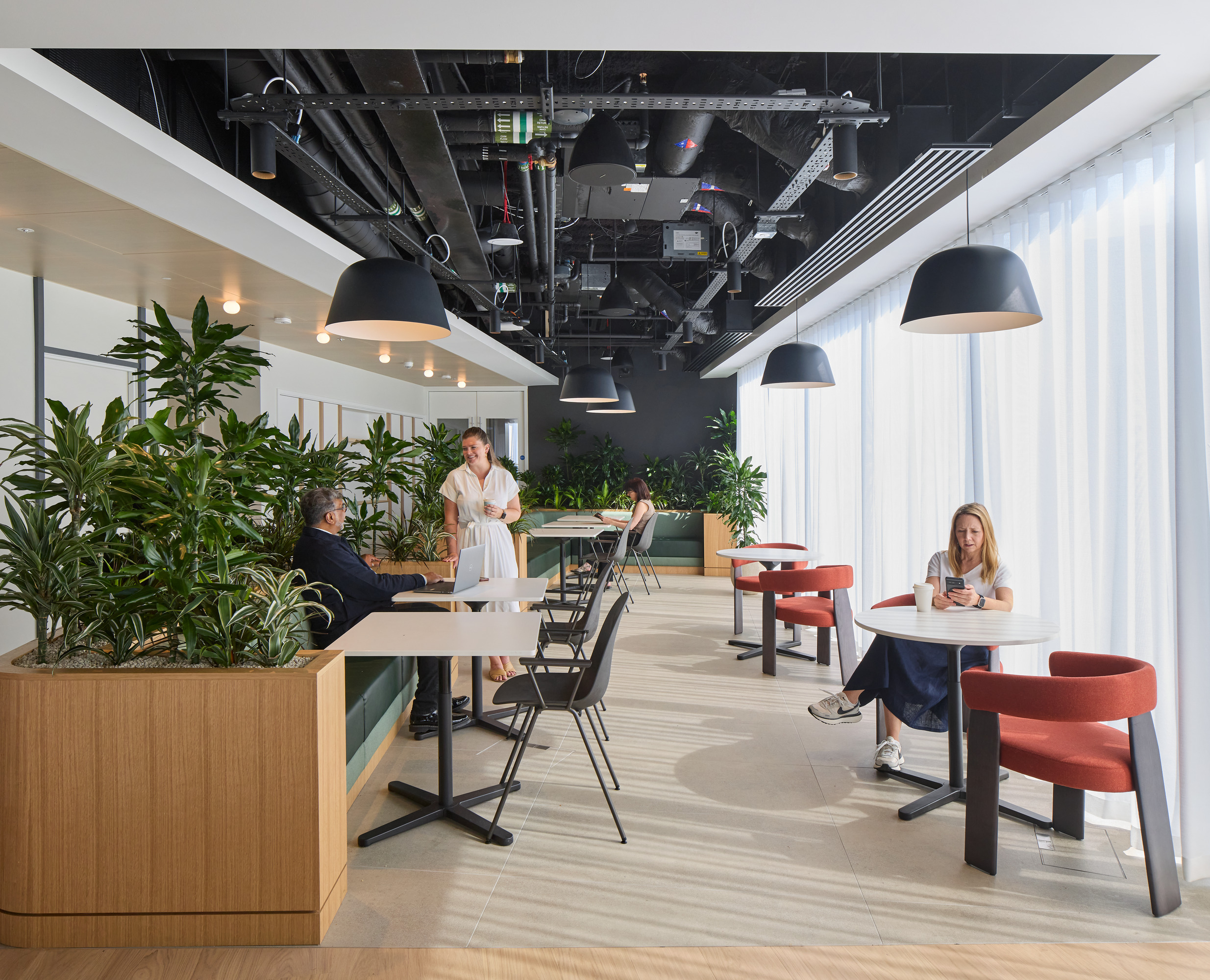 A modern office interior featuring biophilic design elements, with vibrant greenery integrated into wooden planter boxes. The space includes sleek, minimalist furniture: round tables, black chairs, and stylish red upholstered chairs. Ample natural light filters through sheer window treatments, creating an inviting atmosphere for collaboration and relaxation.