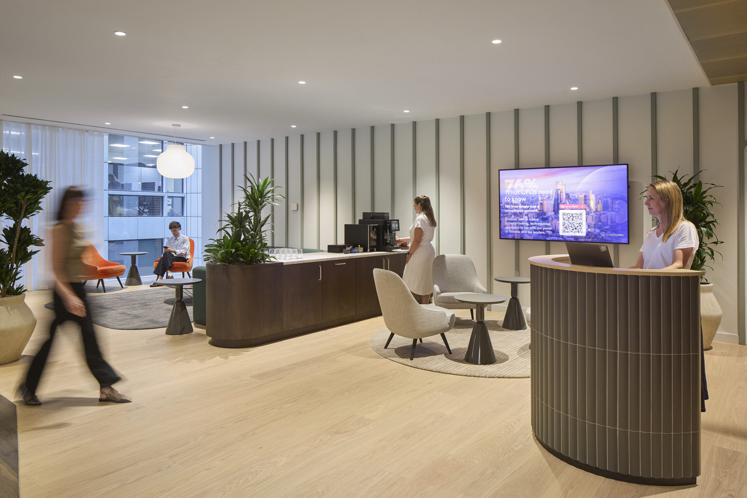 Modern reception area featuring a sleek wooden desk with stylish seating arrangements. The space is enhanced by vertical wall panels in soft hues, abundant greenery, and a large digital display. Natural light floods through expansive windows, creating an inviting atmosphere for visitors and staff alike.