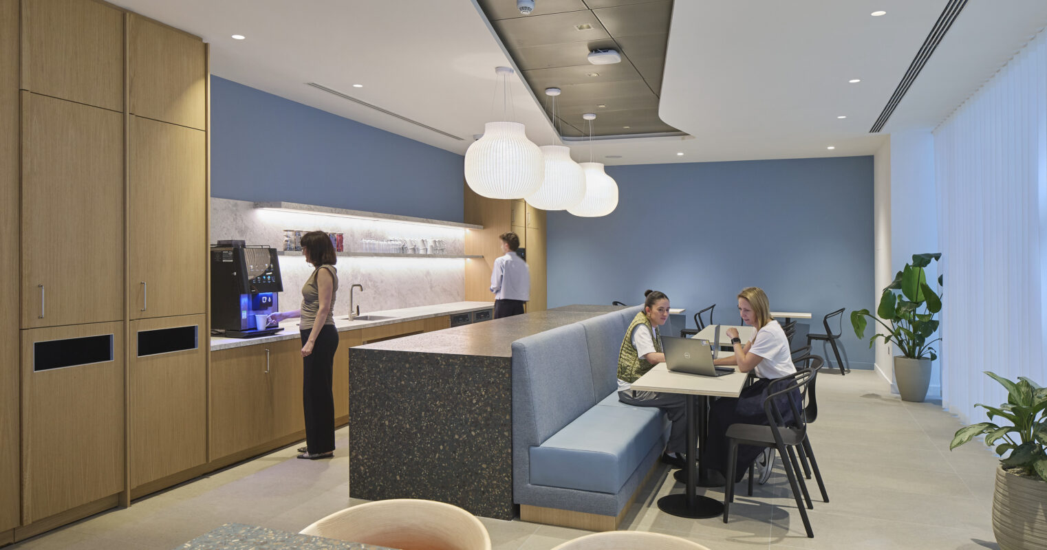 A modern office break room featuring a sleek wood cabinetry and a textured stone countertop. Two employees collaborate at a spacious table while another prepares coffee. Soft pendant lights illuminate the space, complemented by large windows and indoor plants that enhance a calming atmosphere conducive to relaxation and productivity.