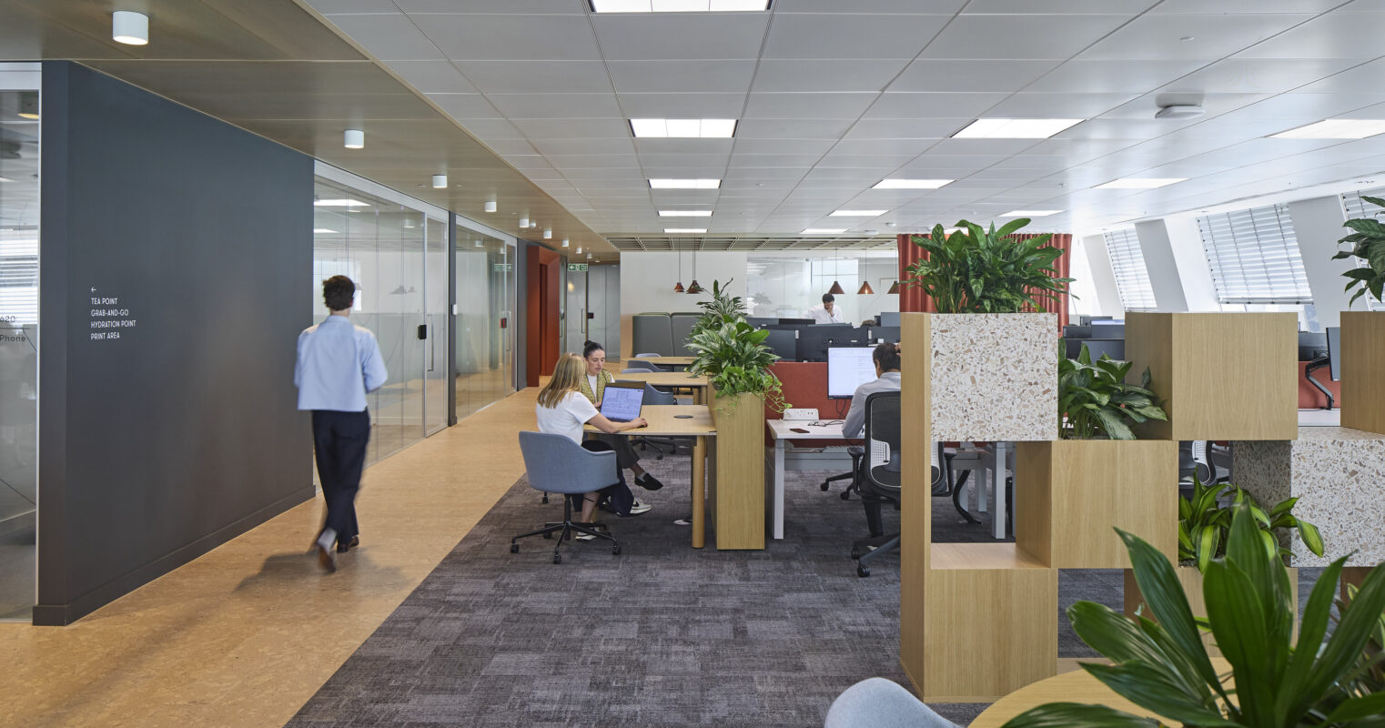 Open-plan office featuring collaborative workspaces with ergonomic desks, modern chairs, and glass partitions. Lush greenery is integrated throughout, providing a refreshing contrast to the contemporary design. Employees engage in various activities, showcasing a blend of functionality and comfort in a well-lit, professional environment.