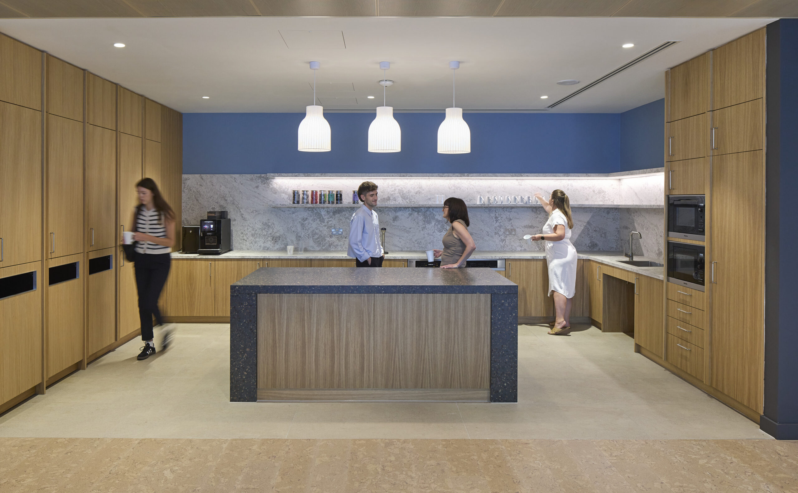 Modern kitchenette featuring warm wooden cabinetry and sleek stone countertops. Pendant lights illuminate the space, highlighting the marble backsplash. Two individuals engage in conversation while another prepares coffee, showcasing a blend of functionality and social interaction in a stylish, open-plan environment.