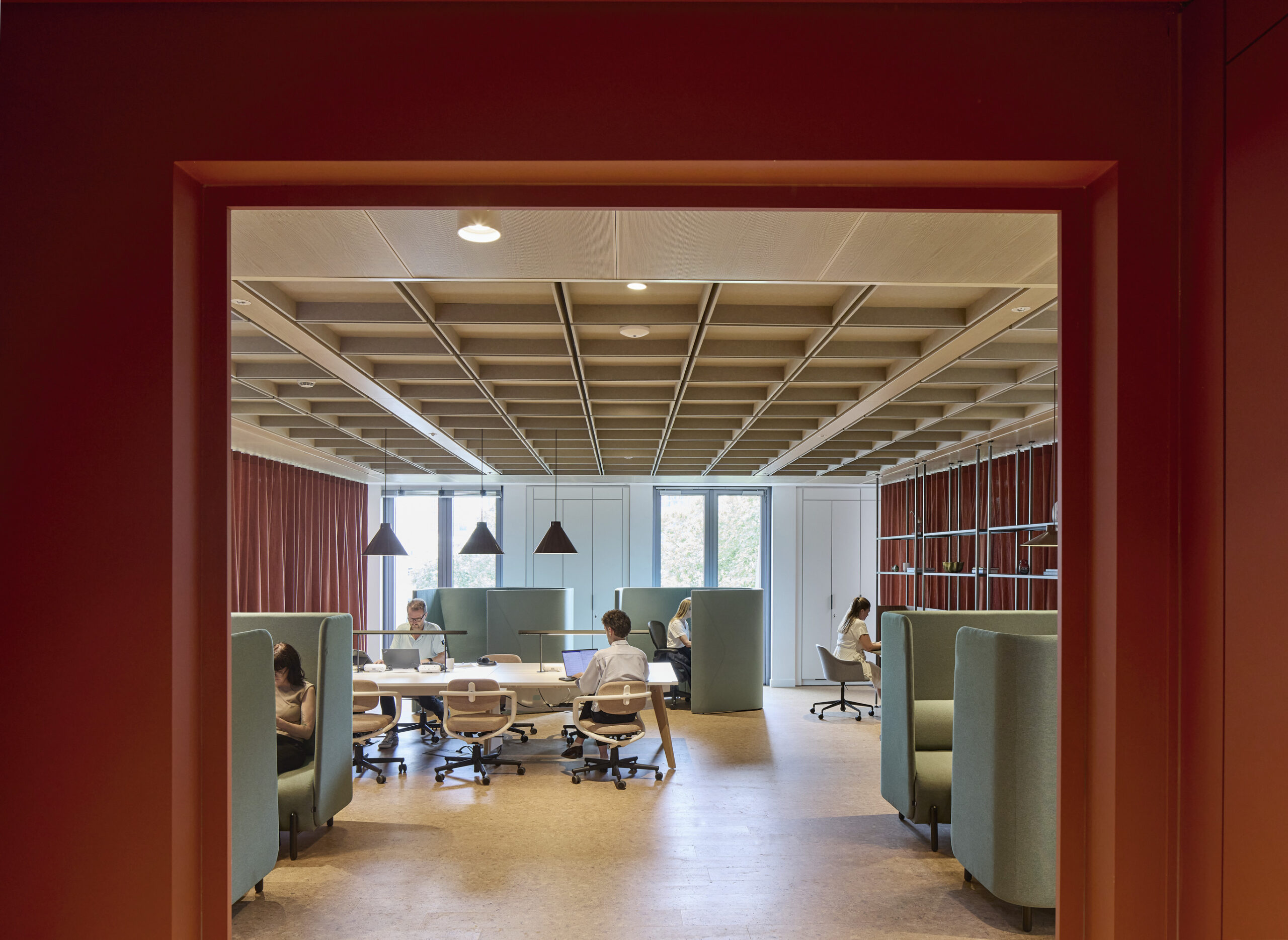 A modern office interior features a spacious layout with collaborative workstations. Ergonomic chairs are paired with sleek tables, while sound-absorbing green booths provide privacy. Natural light floods the room through large windows, complemented by warm wood ceilings and soft pendant lighting, fostering a productive and inviting atmosphere.