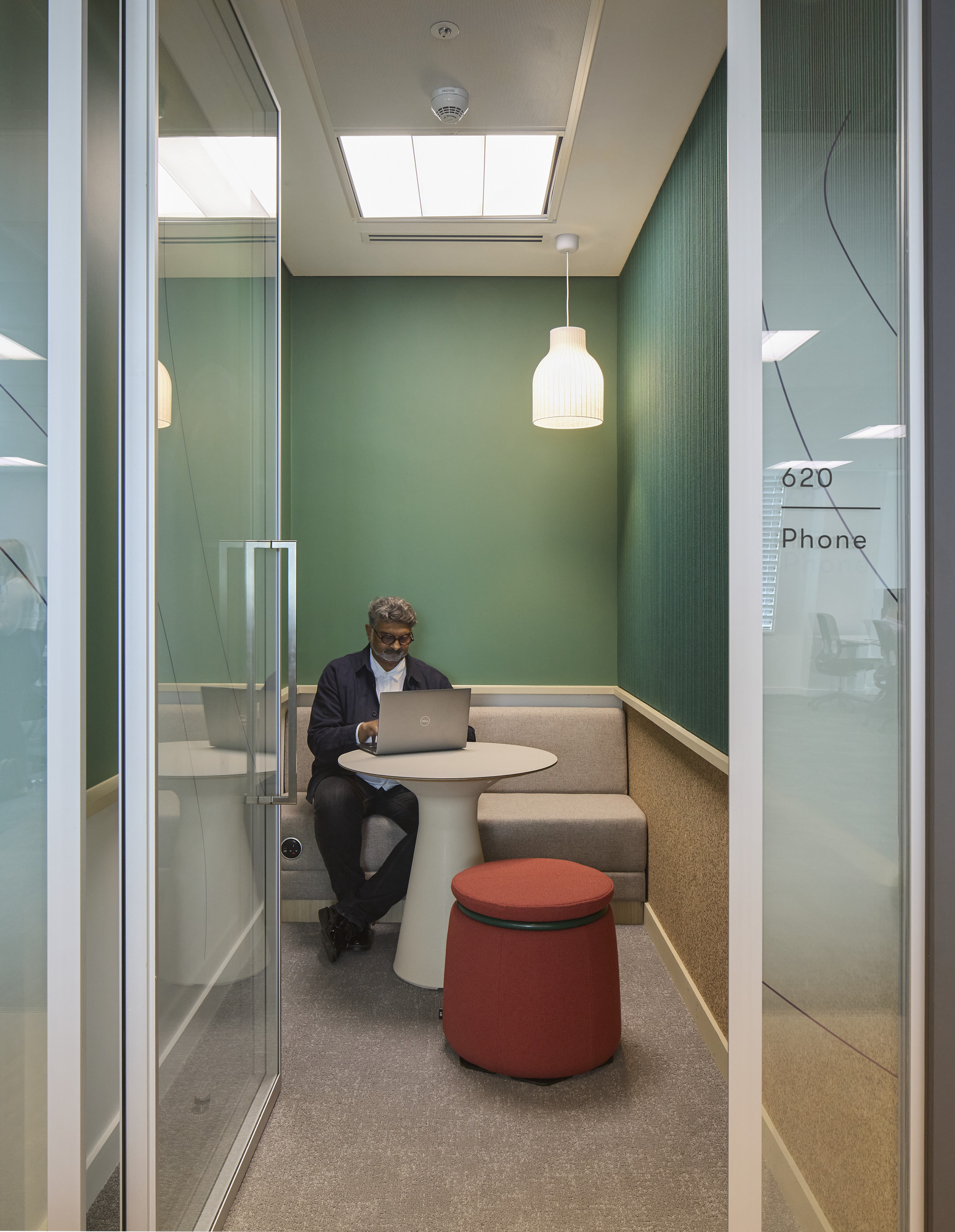 A modern phone booth features a compact round table and a red upholstered stool, fostering a comfortable workspace. The backdrop showcases a vibrant green wall with acoustic panels, promoting sound insulation. Soft pendant lighting enhances the cozy atmosphere, while natural light filters through the ceiling skylight, creating an inviting environment for focused work.