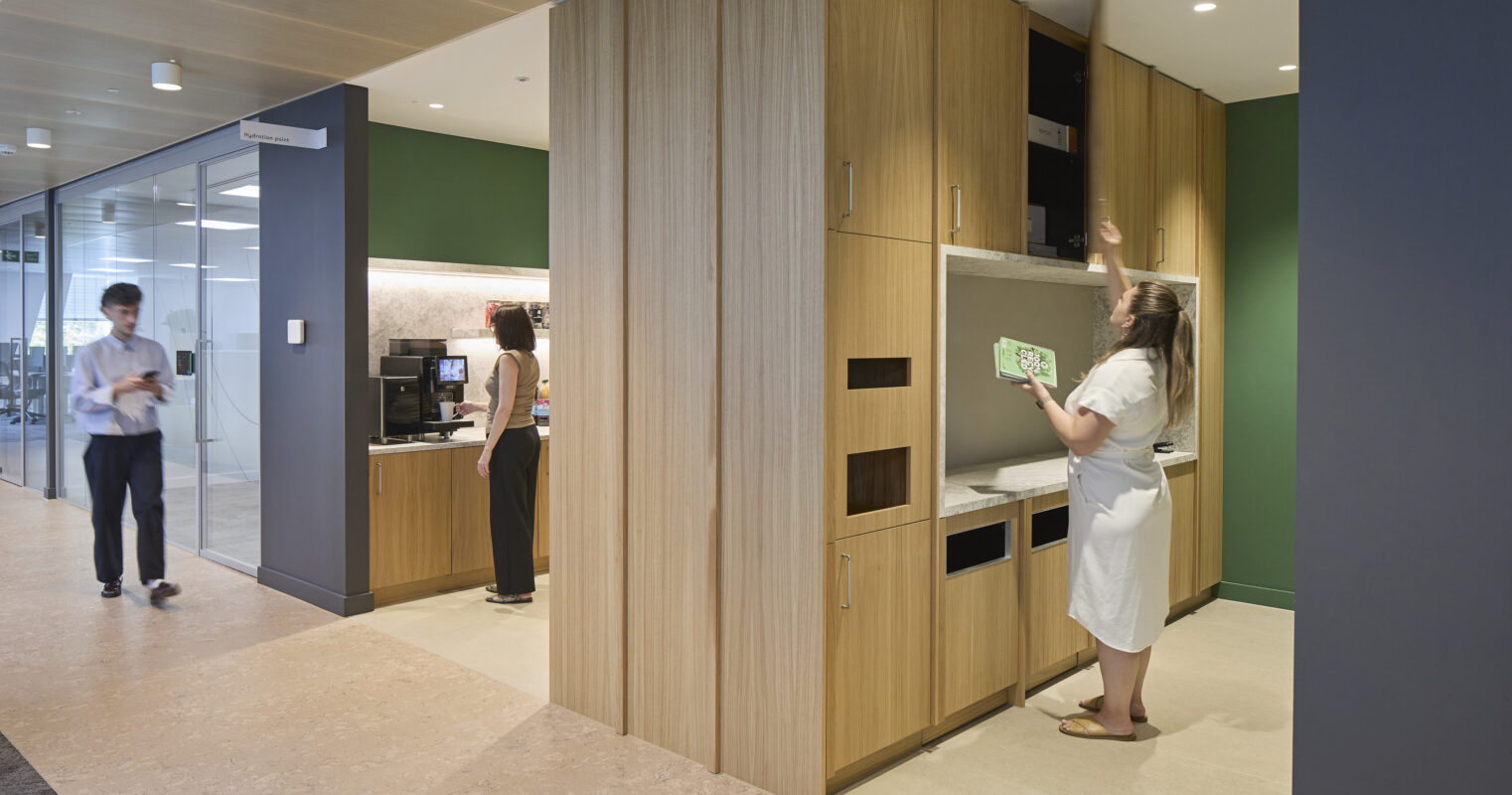 A modern office kitchen features sleek, warm wood cabinetry contrasting with a deep green accent wall. One employee inspects upper storage, while another prepares coffee at a workstation. Natural light filters through glass partitions, enhancing the space's open and collaborative atmosphere. Polished flooring complements the contemporary design.
