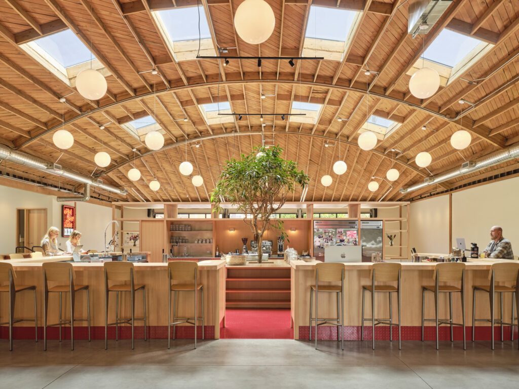 Bright and inviting interior featuring a spacious café with a stunning wood-beamed ceiling and circular skylights. A central potted tree adds a natural element, while warm wooden accents complement the modern bar stools. Elegant pendant lights illuminate the area, creating a welcoming atmosphere for patrons and staff.