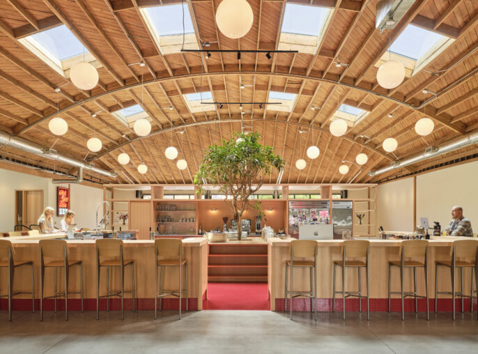 Bright and inviting interior featuring a spacious café with a stunning wood-beamed ceiling and circular skylights. A central potted tree adds a natural element, while warm wooden accents complement the modern bar stools. Elegant pendant lights illuminate the area, creating a welcoming atmosphere for patrons and staff.