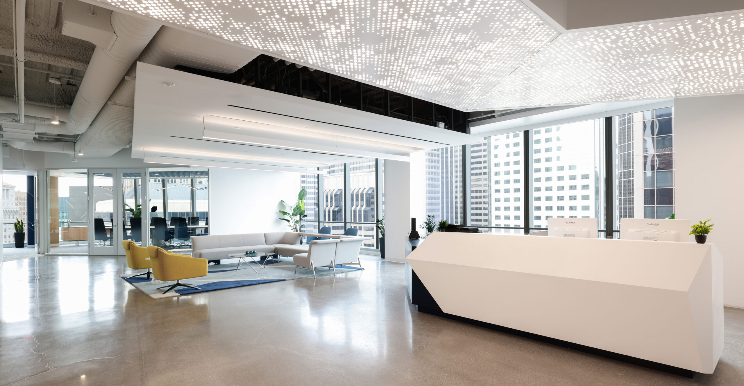 Bright, modern office space featuring a sleek, angular reception desk in white with geometric accents. A contemporary lounge area includes a gray sofa, vibrant yellow chairs, and a patterned area rug. Large windows allow ample natural light, showcasing a city skyline. Minimalist design elements create an airy atmosphere.