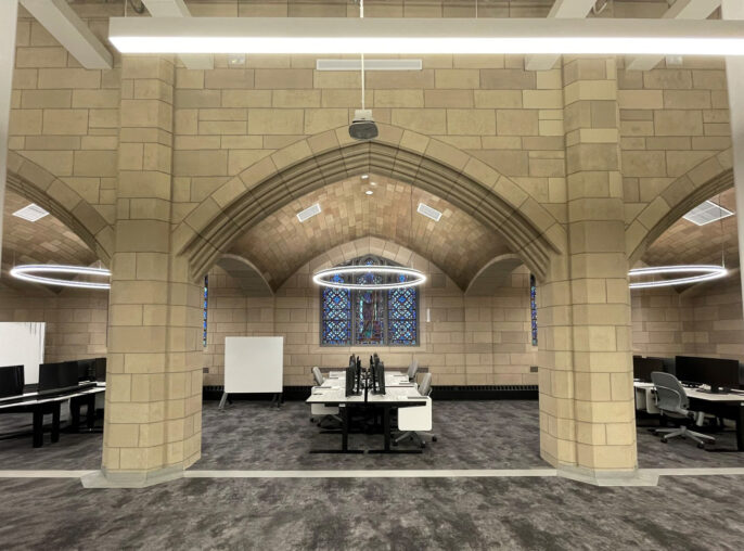Brightly lit workspace features vaulted stone ceilings with intricate archways, blending historic architecture with modern design. Circular LED lighting fixtures hang overhead, illuminating contemporary desks arranged in a collaborative layout. Stained glass windows add a colorful focal point, enhancing the unique character of the environment.