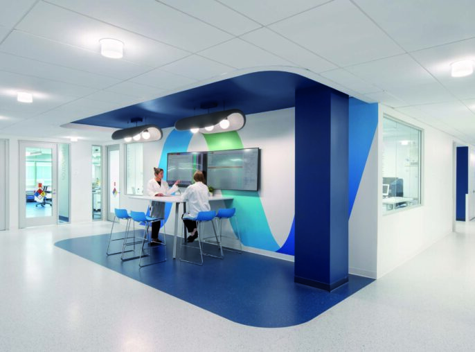 Contemporary healthcare workspace featuring a spacious hallway designed for efficient collaboration. The area showcases a striking blue and green accent wall, complemented by modern circular lighting. Ergonomic seating around a digital display fosters a productive environment for healthcare professionals, enhancing communication and workflow in a clinical setting.
