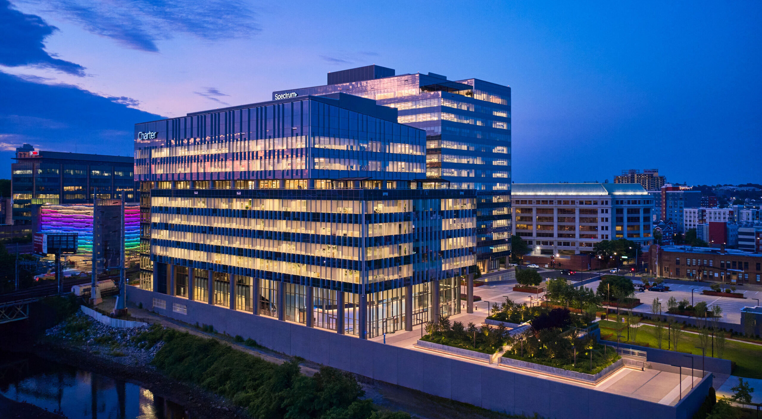 Contemporary office building featuring a striking glass façade that reflects the evening skyline. The sleek design incorporates sustainable materials and expansive windows, maximizing natural light. Surrounding landscaped areas enhance its modern aesthetic, creating an inviting atmosphere for both occupants and visitors in a vibrant urban setting.