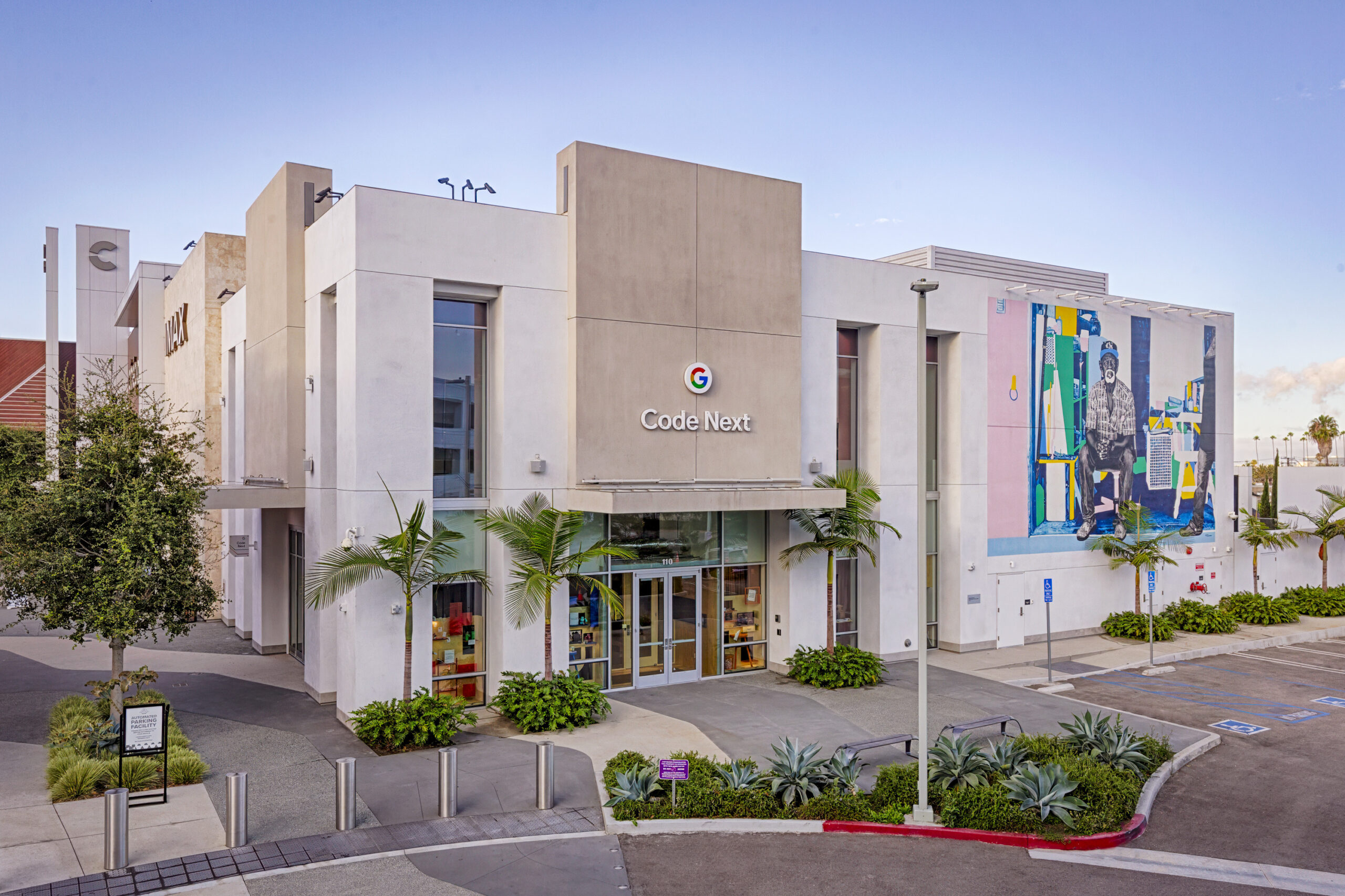The modern facade of the Code Next building features sleek white stucco walls and expansive glass windows. A vibrant mural adorns one side, showcasing colorful figures that enhance the contemporary design. Lush landscaping, including palm trees and succulents, complements the urban environment, creating an inviting atmosphere.