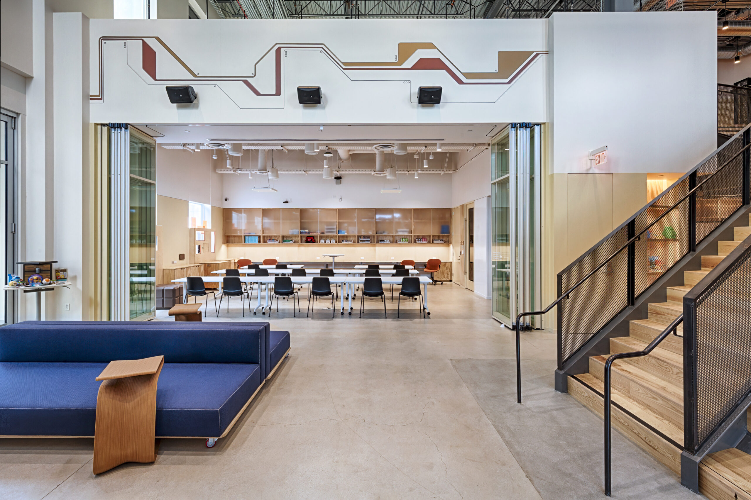 Contemporary open-plan workspace featuring a blend of natural wood and modern materials. A sleek blue sofa complements the minimalist design. The central area showcases a large conference table surrounded by black chairs, framed by glass partitions, enhancing collaboration and transparency within the environment. Warm lighting highlights the space's inviting atmosphere.