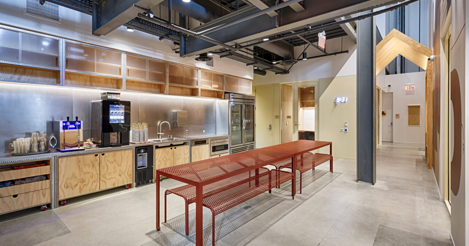 A modern industrial kitchen featuring sleek stainless steel appliances, wooden cabinetry, and an open layout. A bold red metal dining table complements the space, surrounded by minimalist design elements. Large windows provide natural light, enhancing the contemporary atmosphere and encouraging collaboration.