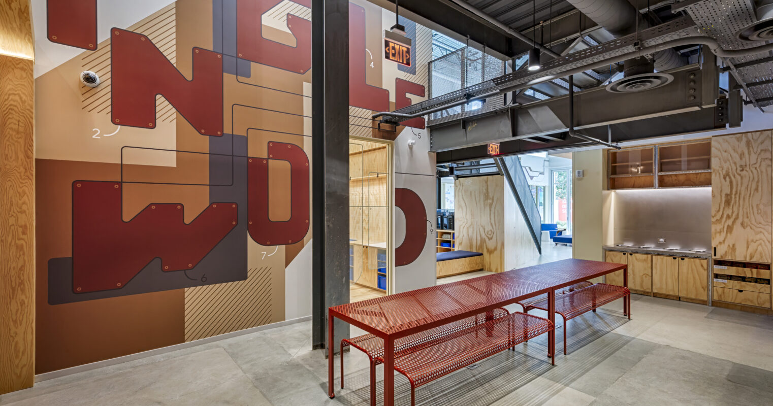 A modern collaborative workspace featuring a striking mural with bold, geometric shapes in deep reds and browns. Industrial elements include exposed steel beams and ductwork, complementing the warm wood accents. A sleek, red metal table encourages group interaction, enhancing the open design's functionality and creativity.