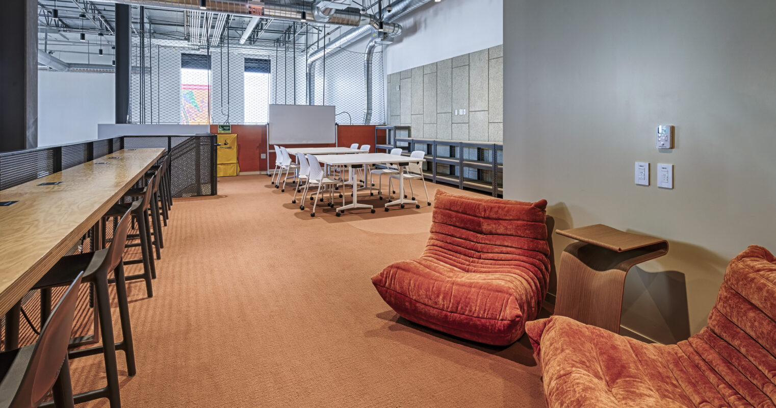 Brightly lit collaborative workspace featuring cozy, vibrant orange seating alongside a long wooden table with ergonomic stools. The backdrop includes soft gray acoustic panels and modular white furniture, promoting a modern and inviting atmosphere for creativity and teamwork. Textured carpets enhance comfort while maintaining a professional aesthetic.