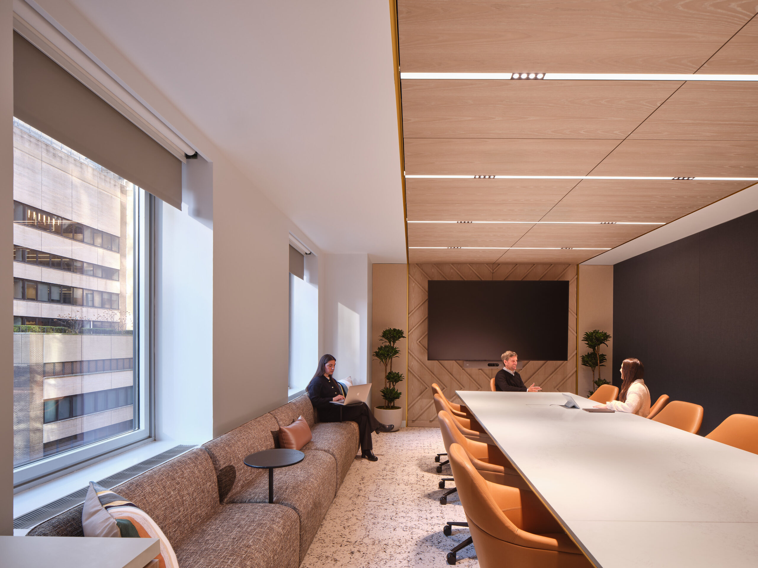 A modern conference room featuring a sleek, elongated table surrounded by ergonomic orange chairs. Natural light floods through large windows, highlighting the warm wood paneling and geometric accent wall. Plush seating offers a cozy corner for collaboration, complemented by greenery that adds a touch of vibrancy to the space.