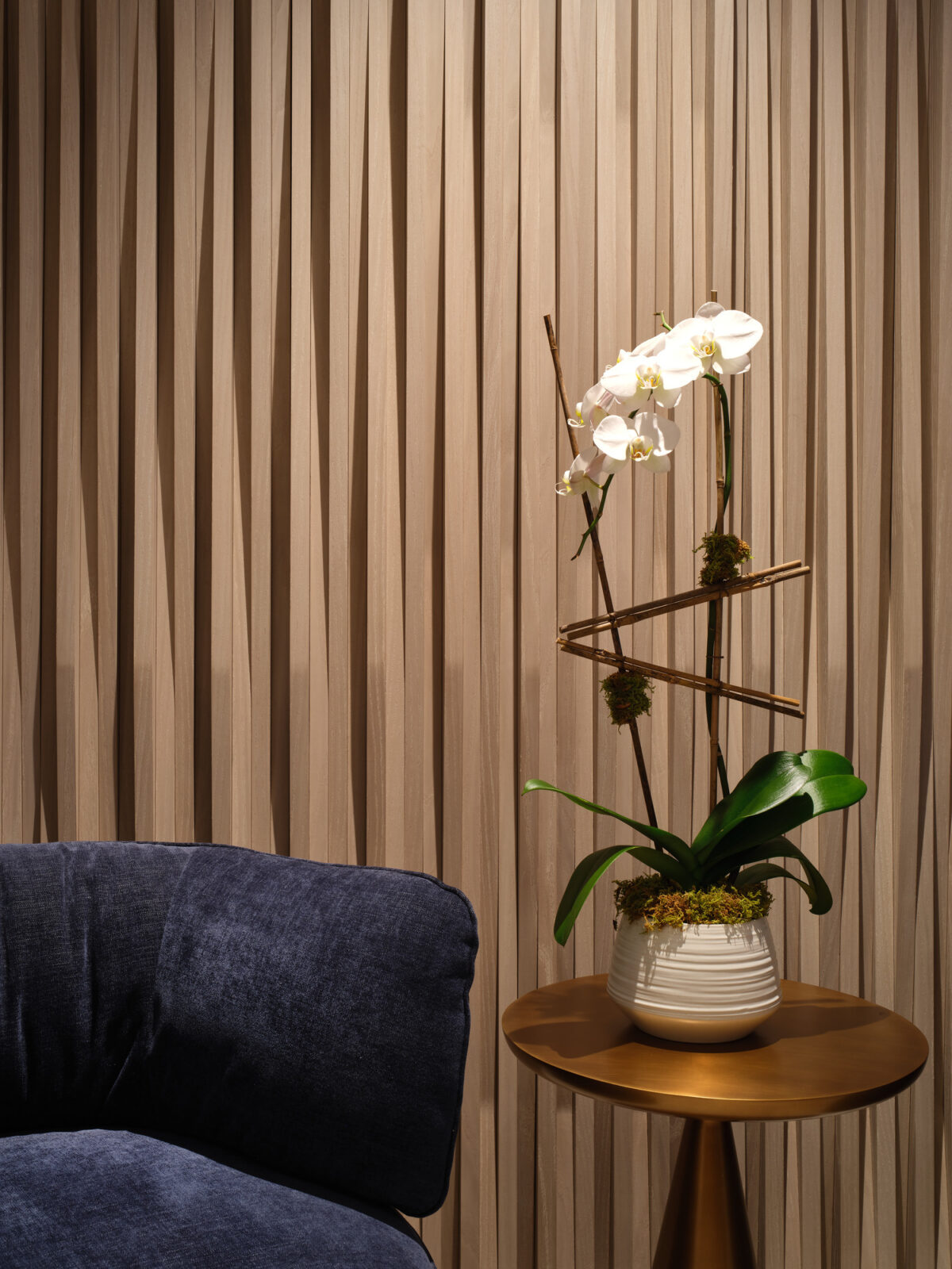 A textured wall featuring vertical wooden slats creates a warm backdrop for a contemporary arrangement. A sleek side table holds a striking orchid, artistically arranged to spiral upward with natural elements. The rich blue upholstery of the lounge chair adds depth, harmonizing with the earthy tones of the design.